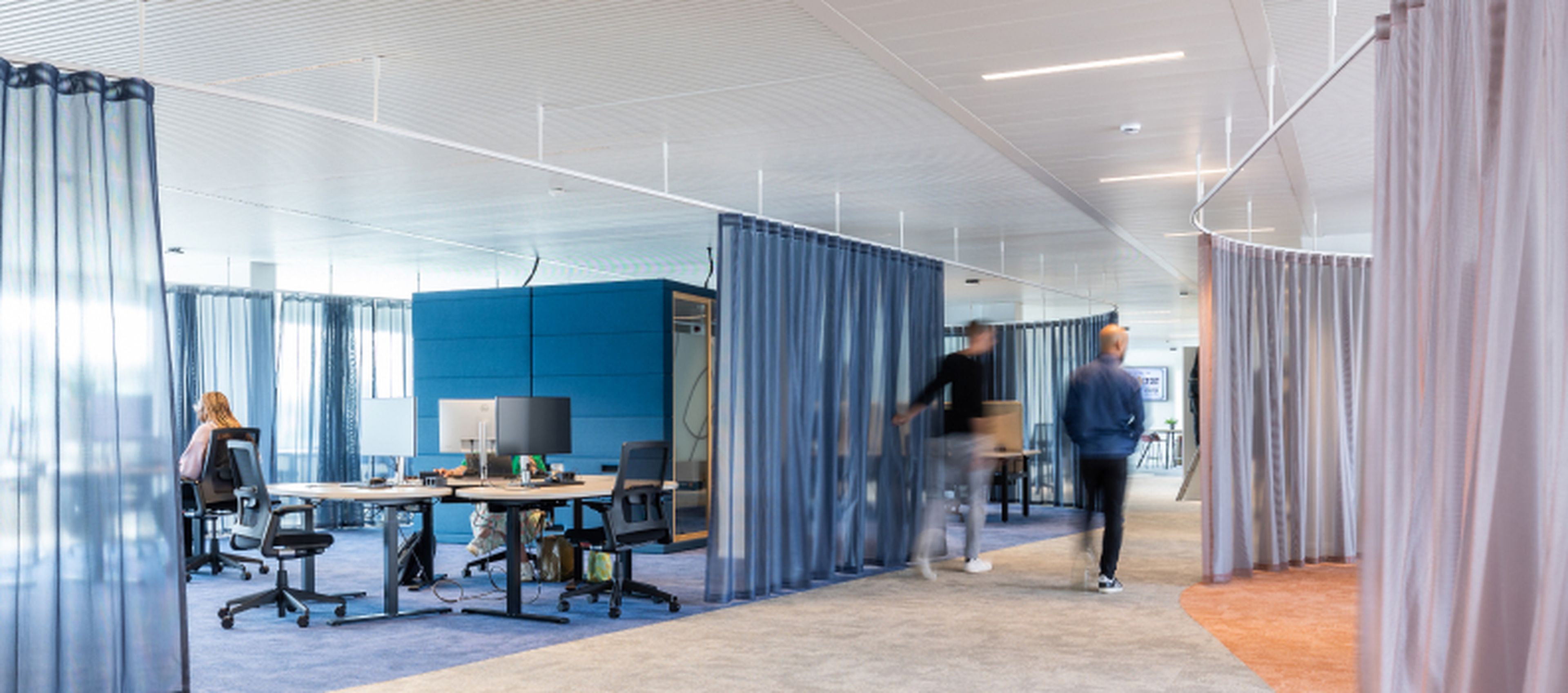 Open kantoorruimte in het hoofdkantoor van Q8 in Antwerpen. Verschillende medewerkers werken of lopen door de moderne en open omgeving, die is opgedeeld met semi-transparante gordijnen in blauw- en grijstinten.