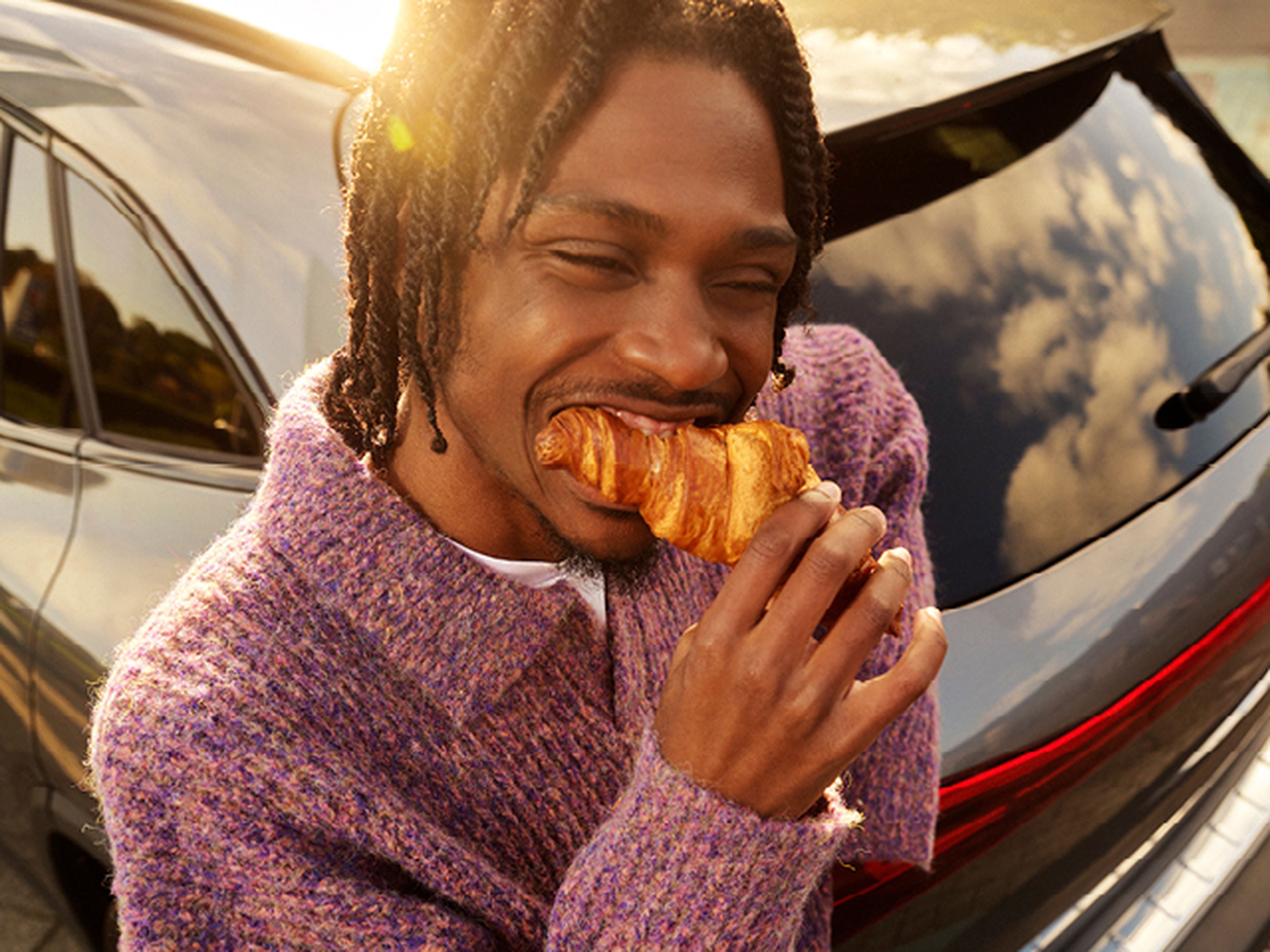 Persoon eet een croissant terwijl hij voor een auto staat.