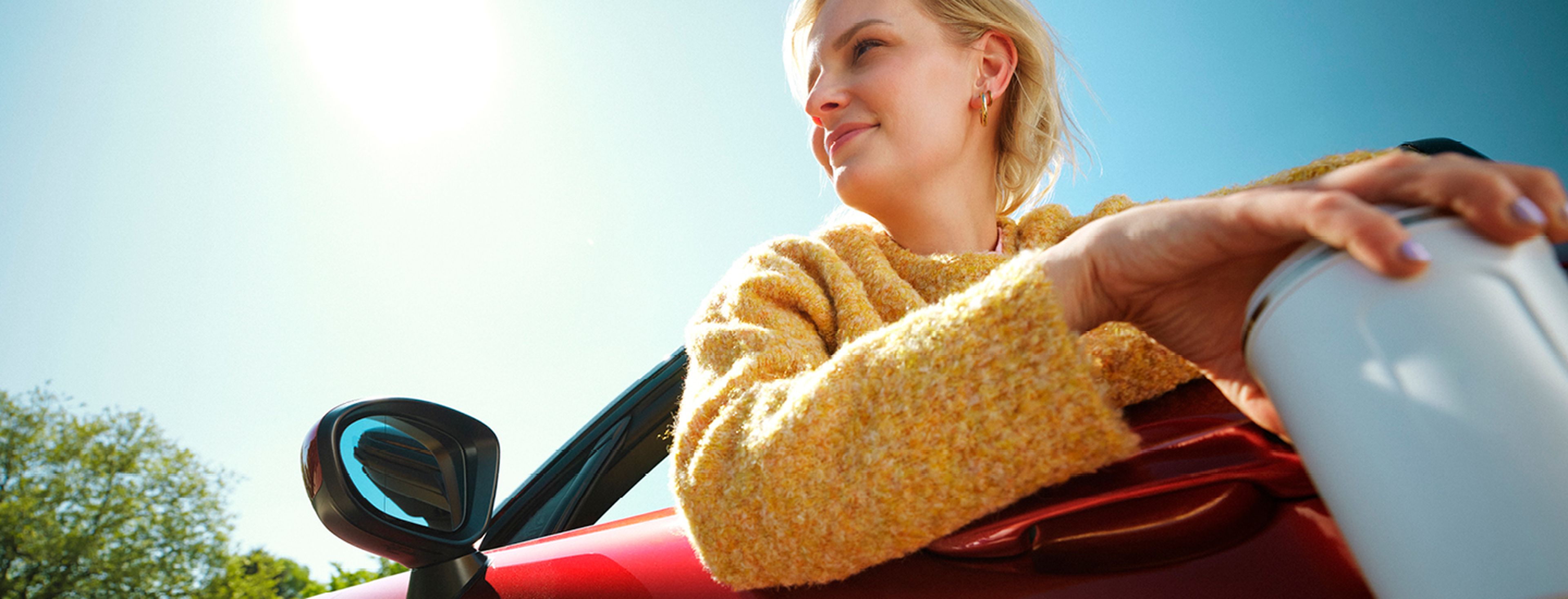 Een jonge vrouw in een gele trui zit ontspannen in een rode cabriolet met haar arm over de deur en een koffiebeker in de hand.