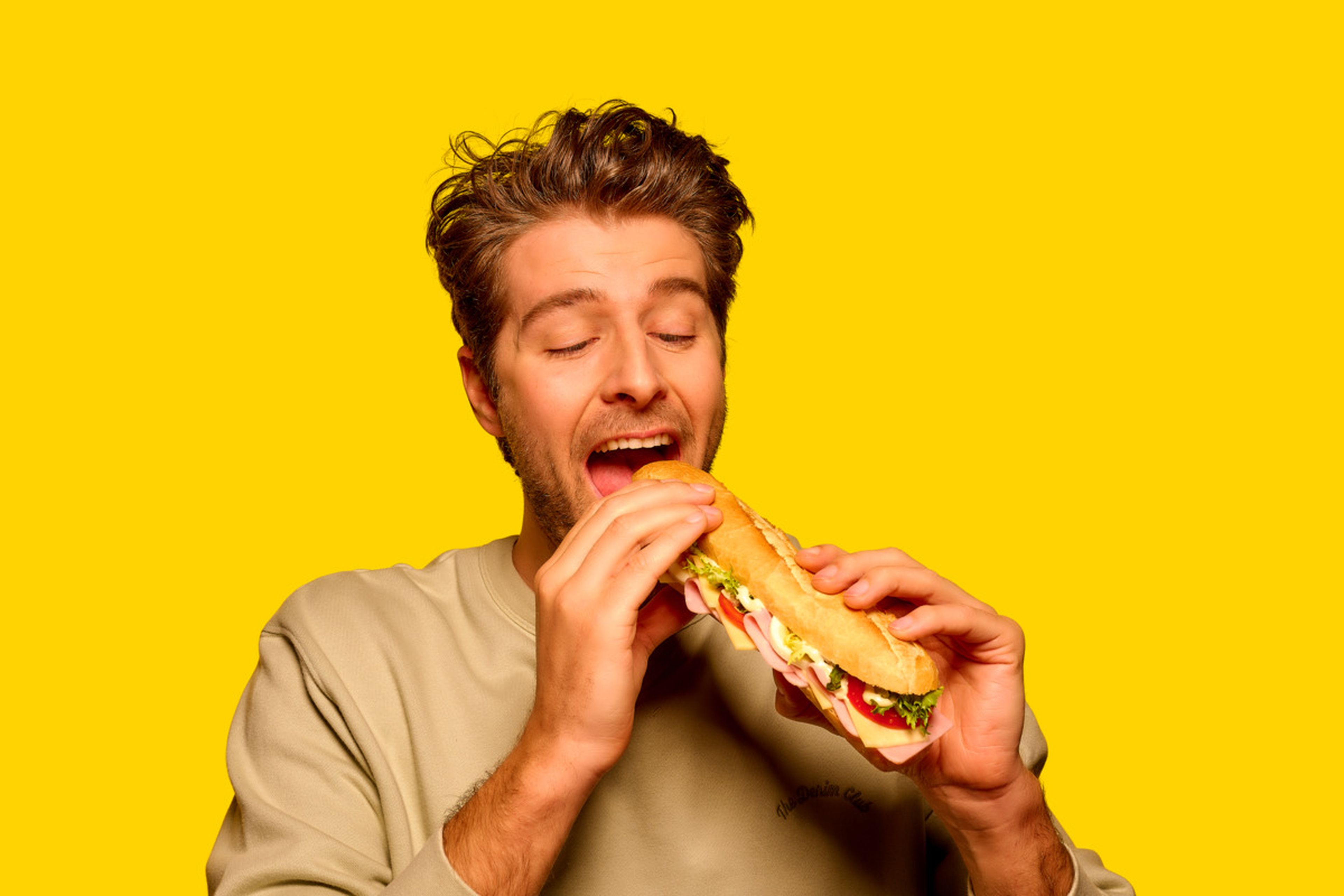 Man met lichtbruin haar in een beige trui staat voor een felgele achtergrond en neemt met gesloten ogen een grote hap van een belegd stokbrood.