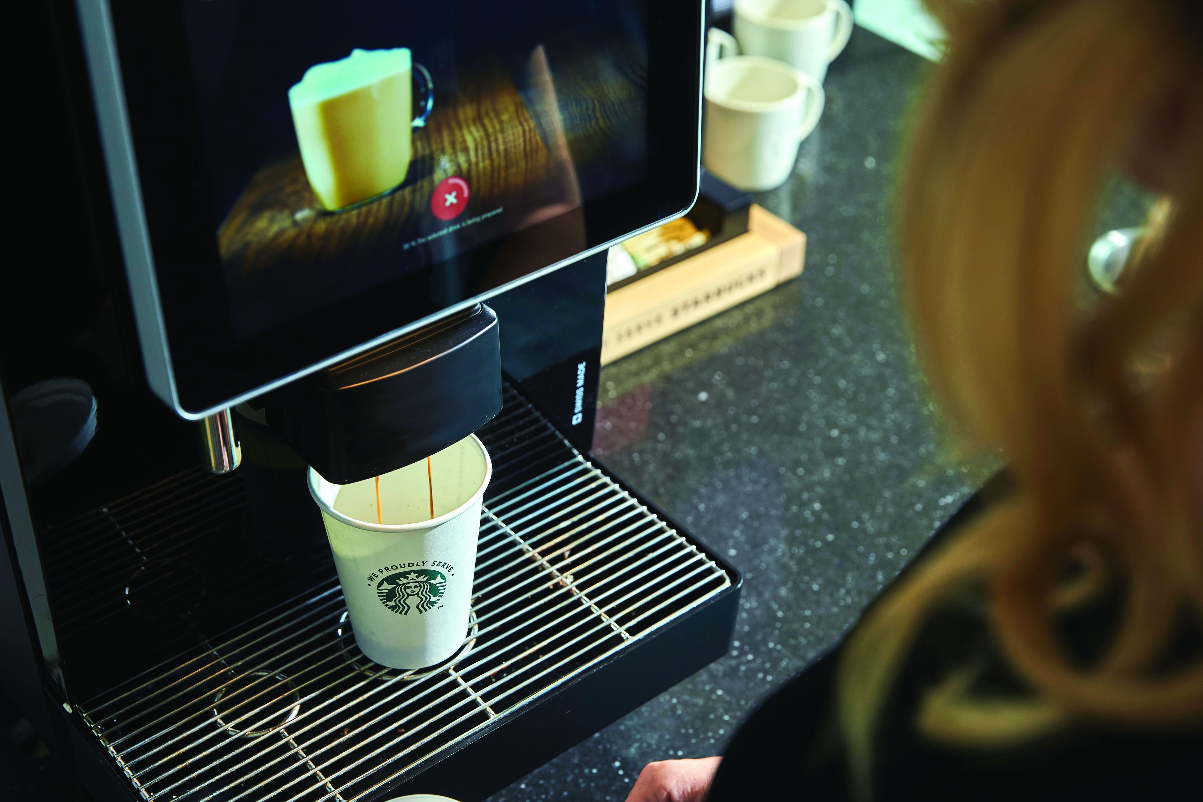 Starbucks coffee machine preparing a hot drink in a paper cup, with touchscreen and a person in the background at a Q8 station.