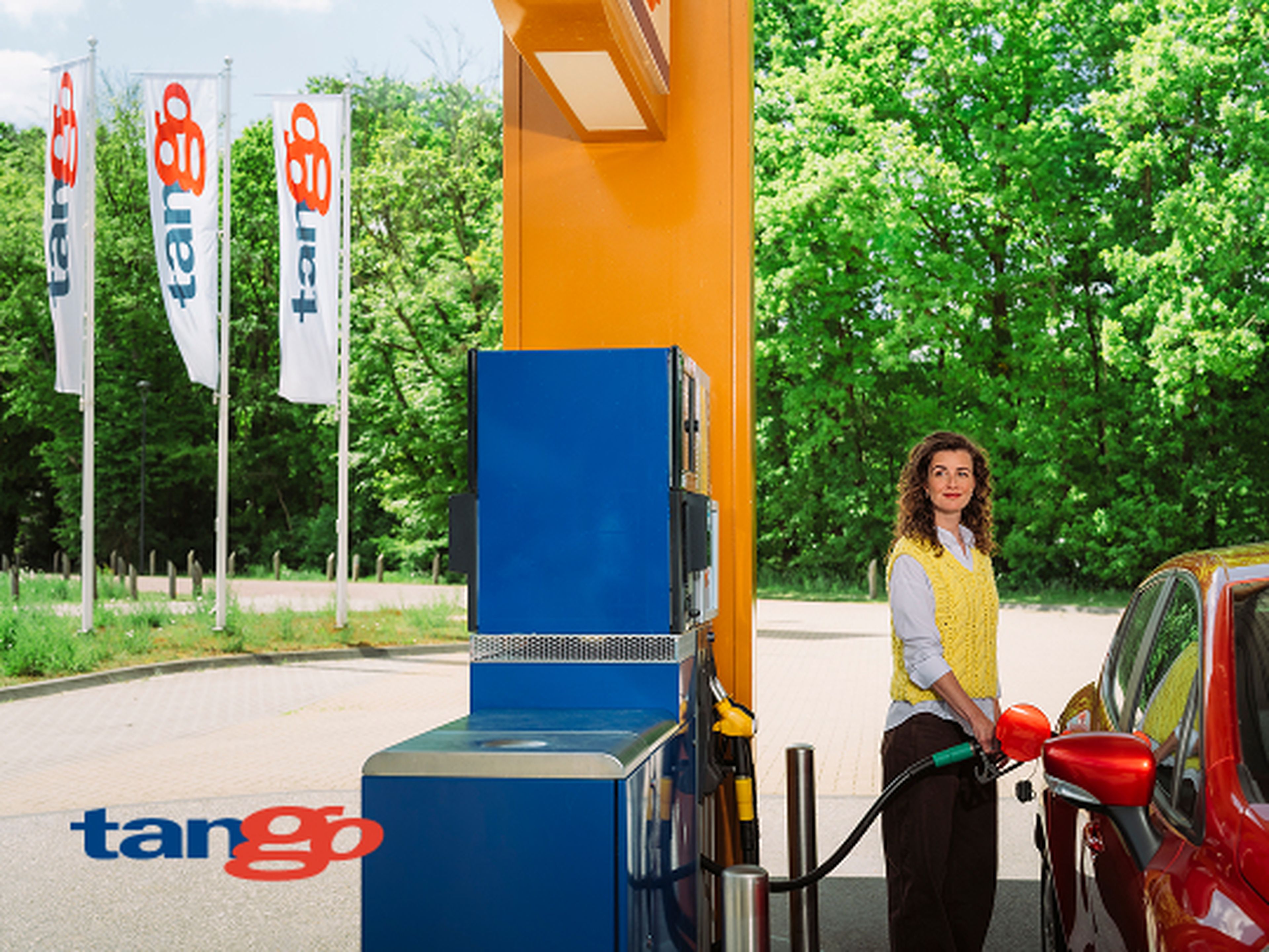 Een vrouw tankt haar rode auto bij een Tango tankstation. Op de achtergrond zijn groene bomen en wapperende Tango-vlaggen zichtbaar.