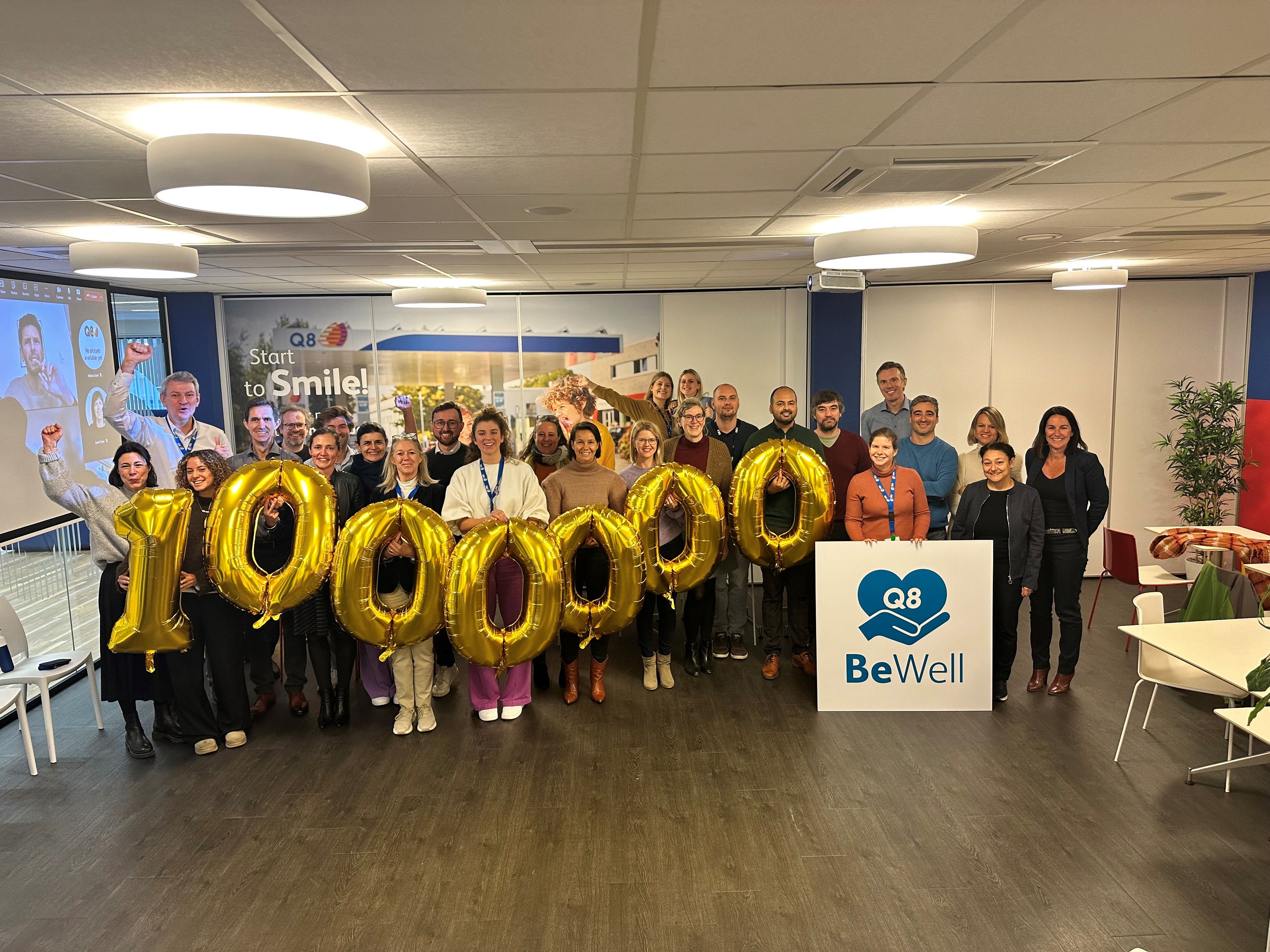 A large group of colleagues is holding numbered balloons, with a BeWell sign in front of them.