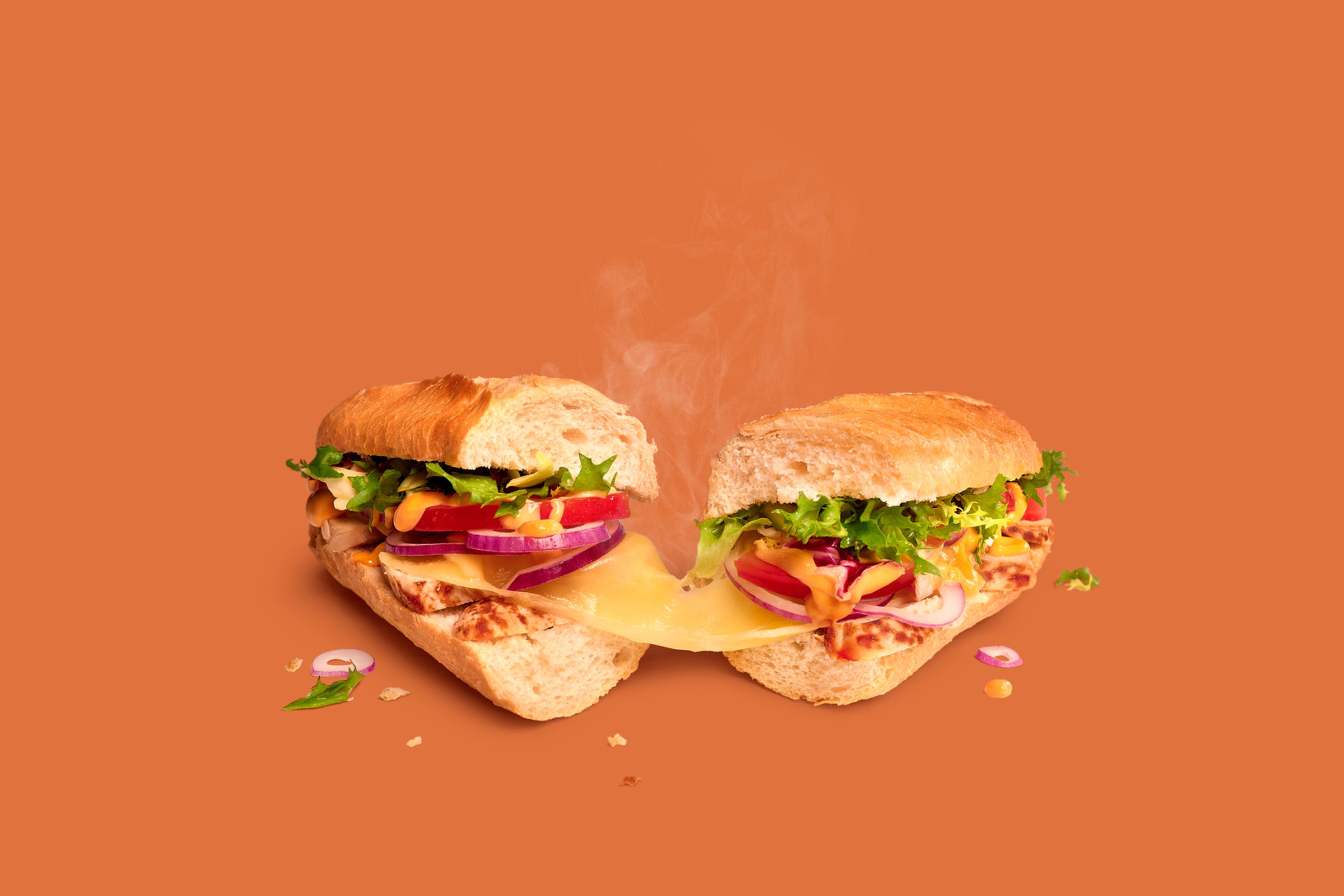 Sandwich Chicken Samouraï Remix de Panos, coupé en deux, avec poulet, fromage, légumes et sauce sur fond orange.