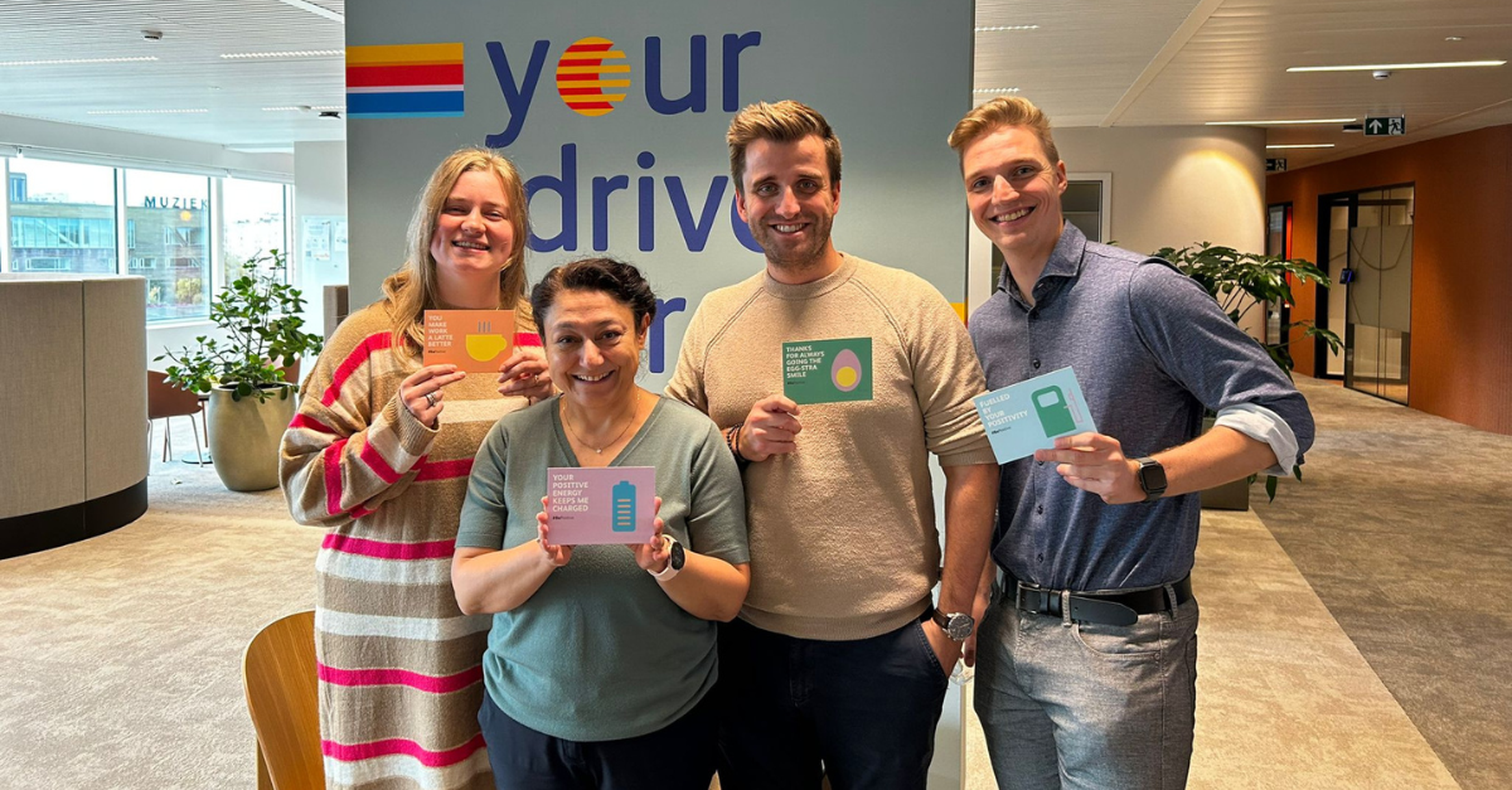 A group of 4 colleagues is smiling at the camera while holding a #BePositive card.