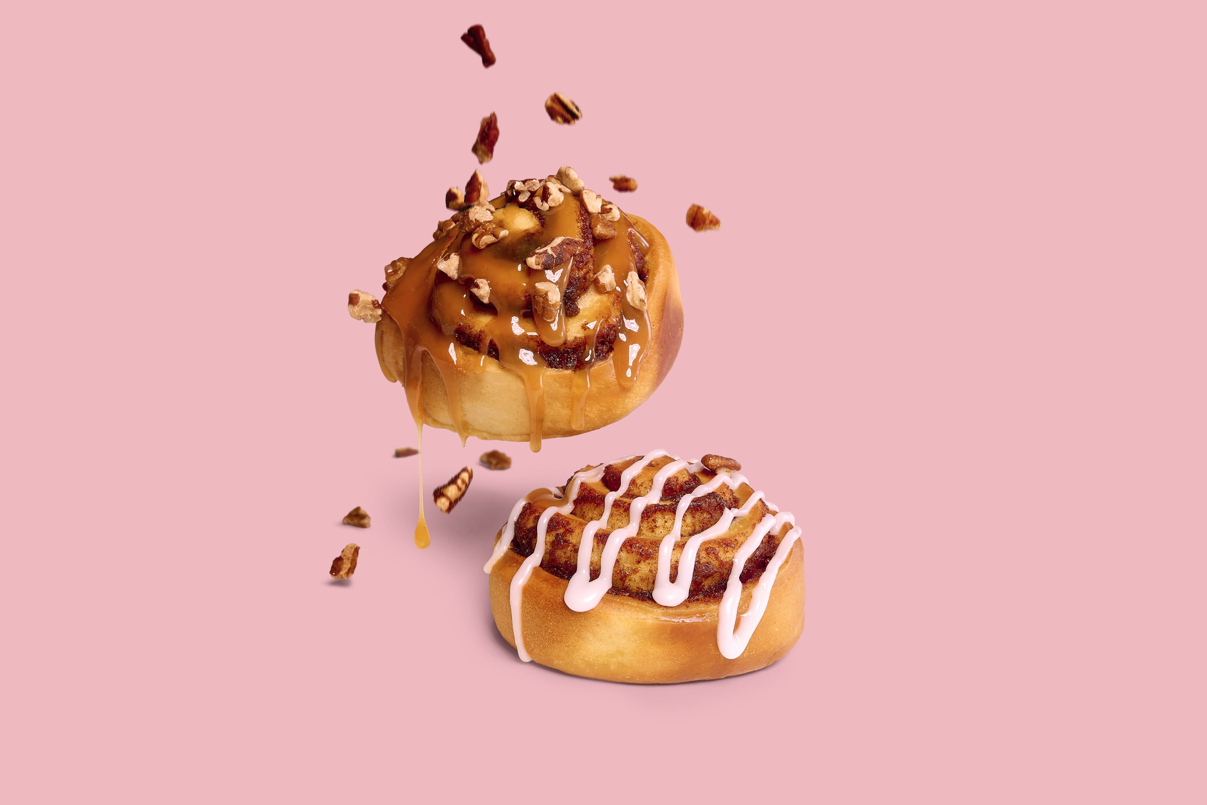 Deux roulés à la cannelle de Panos sur fond rose, avec caramel, noix de pécan et glaçage blanc.