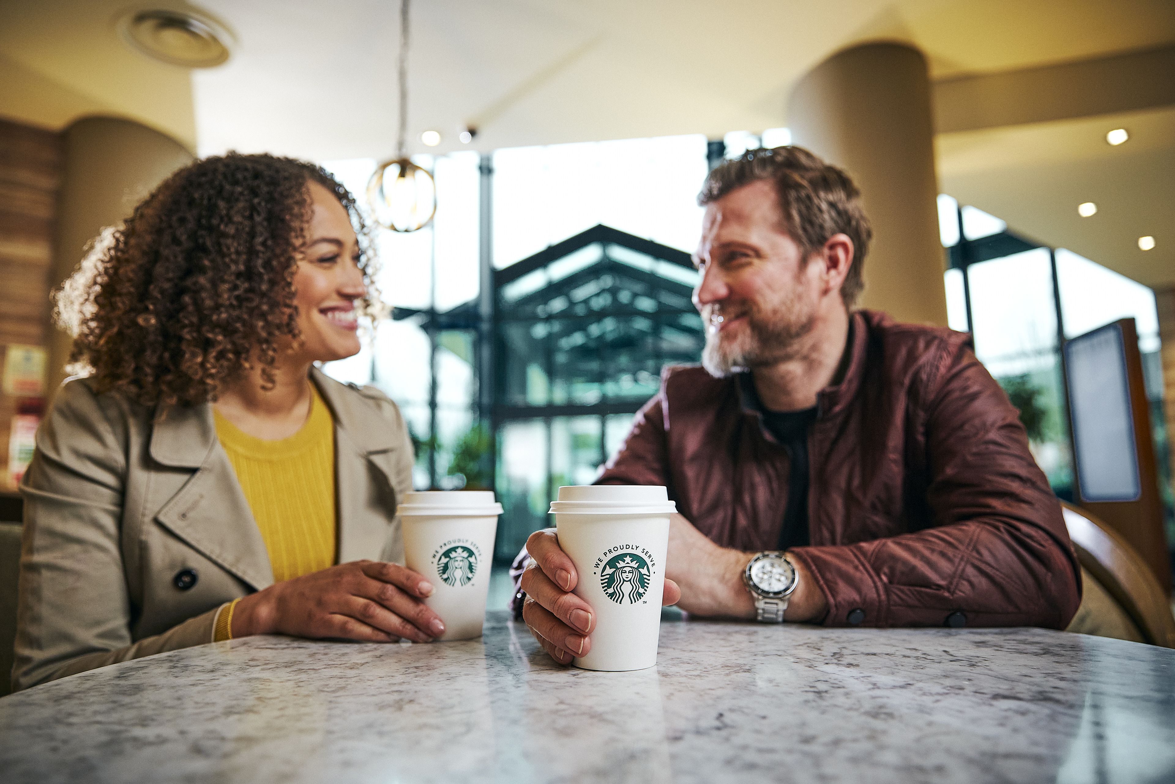 Twee personen zitten aan een tafel met Starbucks-koffiebekers en praten met elkaar.
