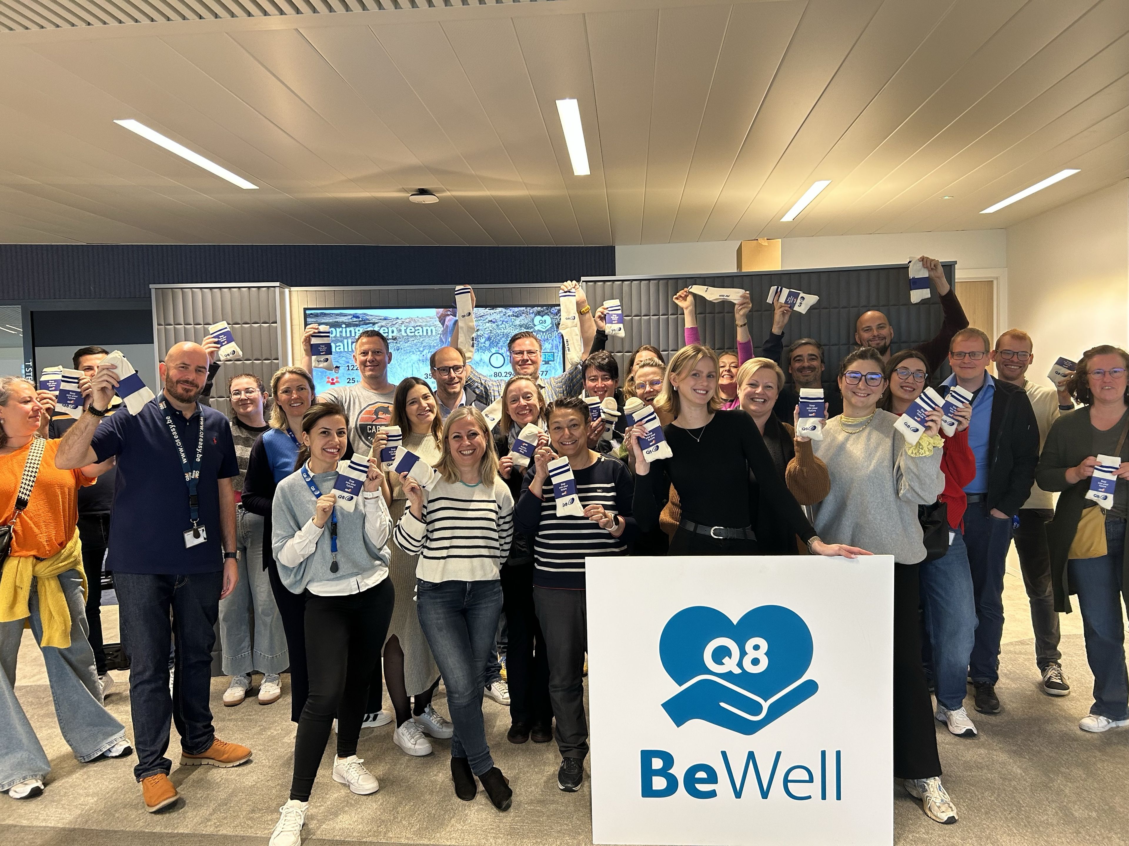 A large group of Q8 colleagues is smiling at the camera and taking a group photo with a BeWell sign in front of them.