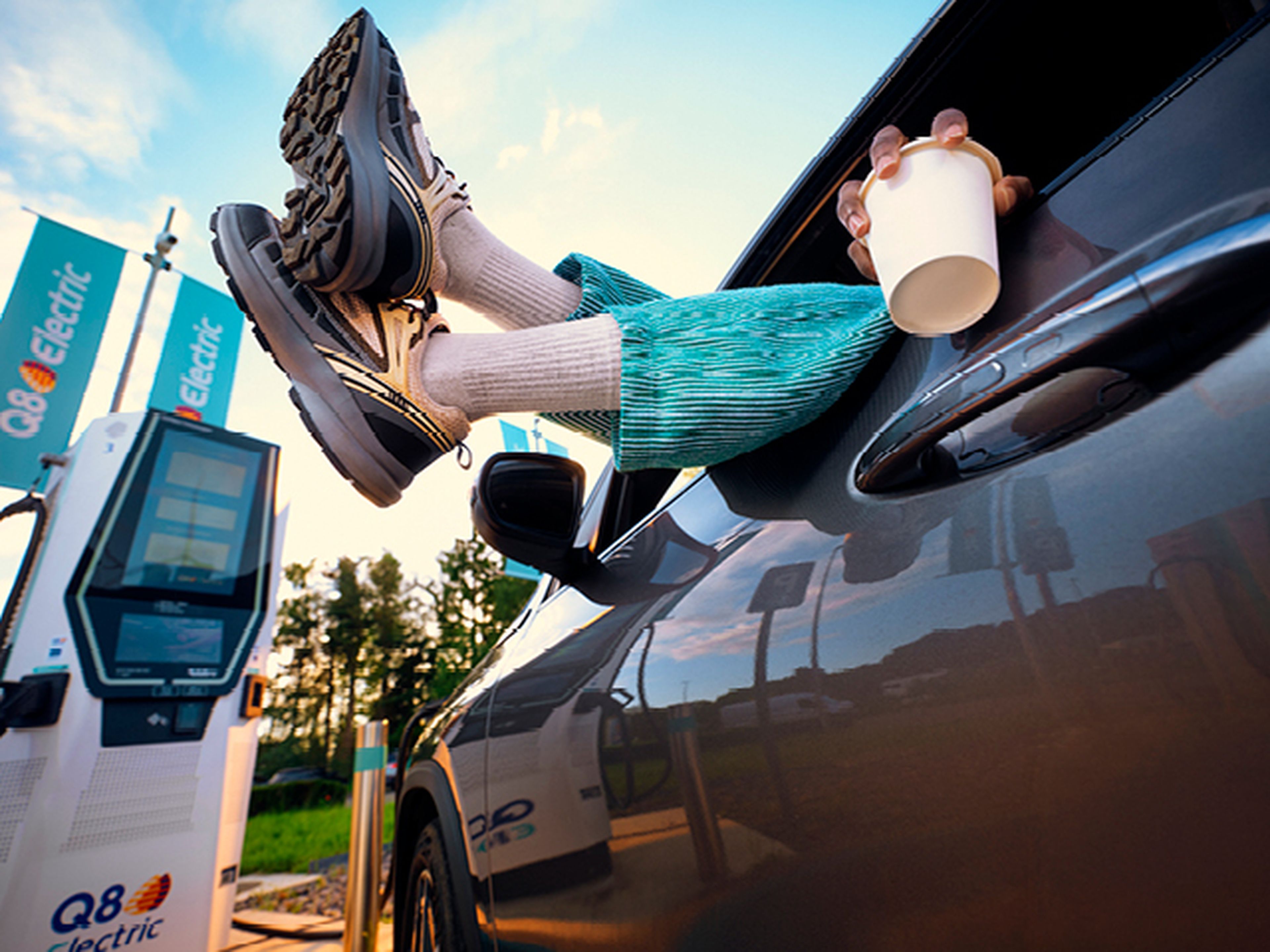 Pieds sortant de la fenêtre de voiture, personne boit du café à la station de recharge Q8 Electric.