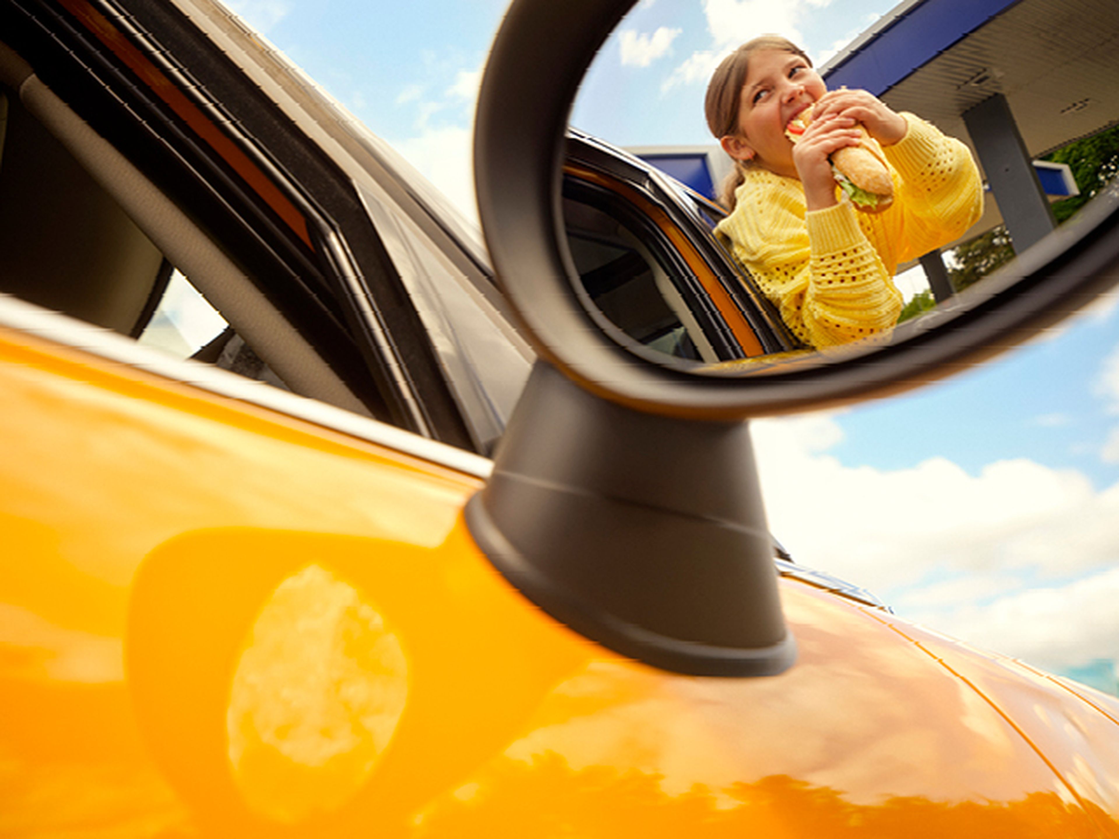Girl eating a sandwich, seen in the side mirror of a car at a Q8 fuel station.
