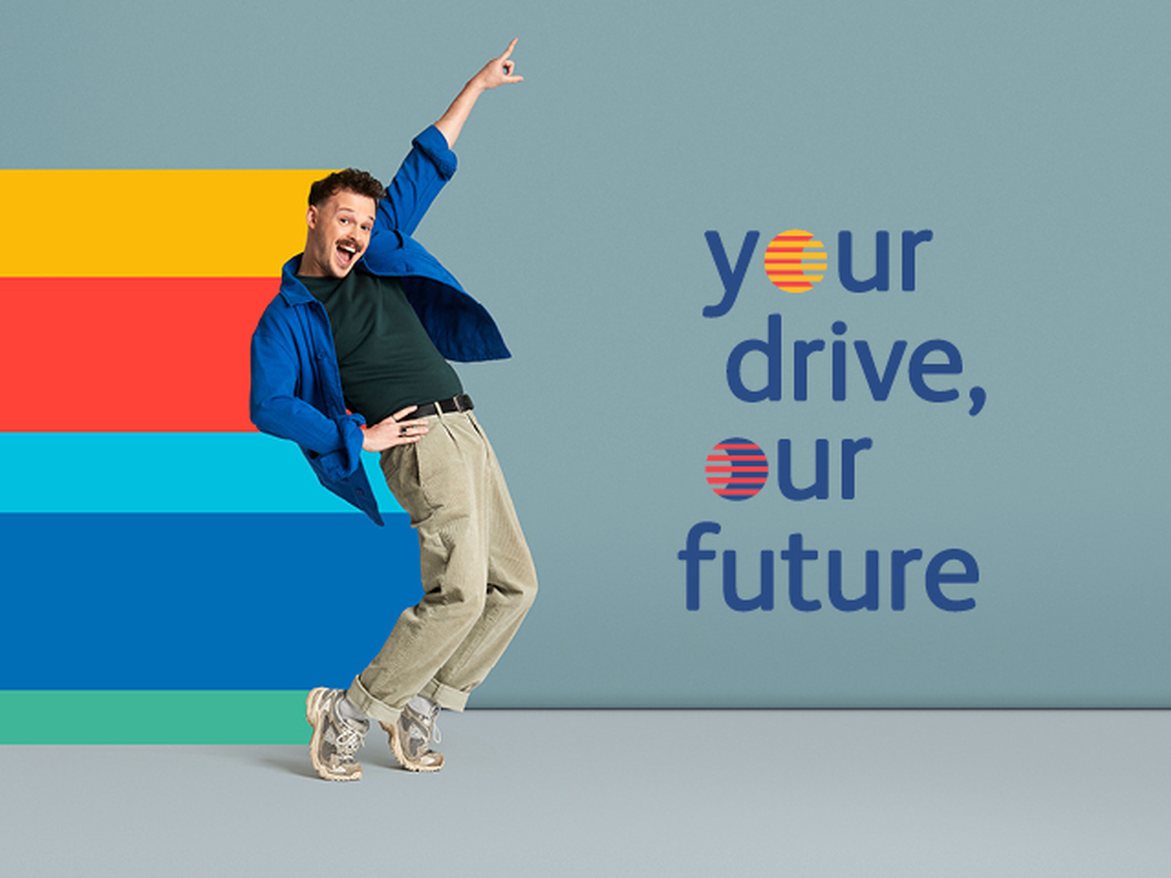 Een persoon in een expressieve pose staat voor een effen blauwe achtergrond en draagt een felblauw overhemd, een donkergroen T-shirt, beige broek en sneakers.