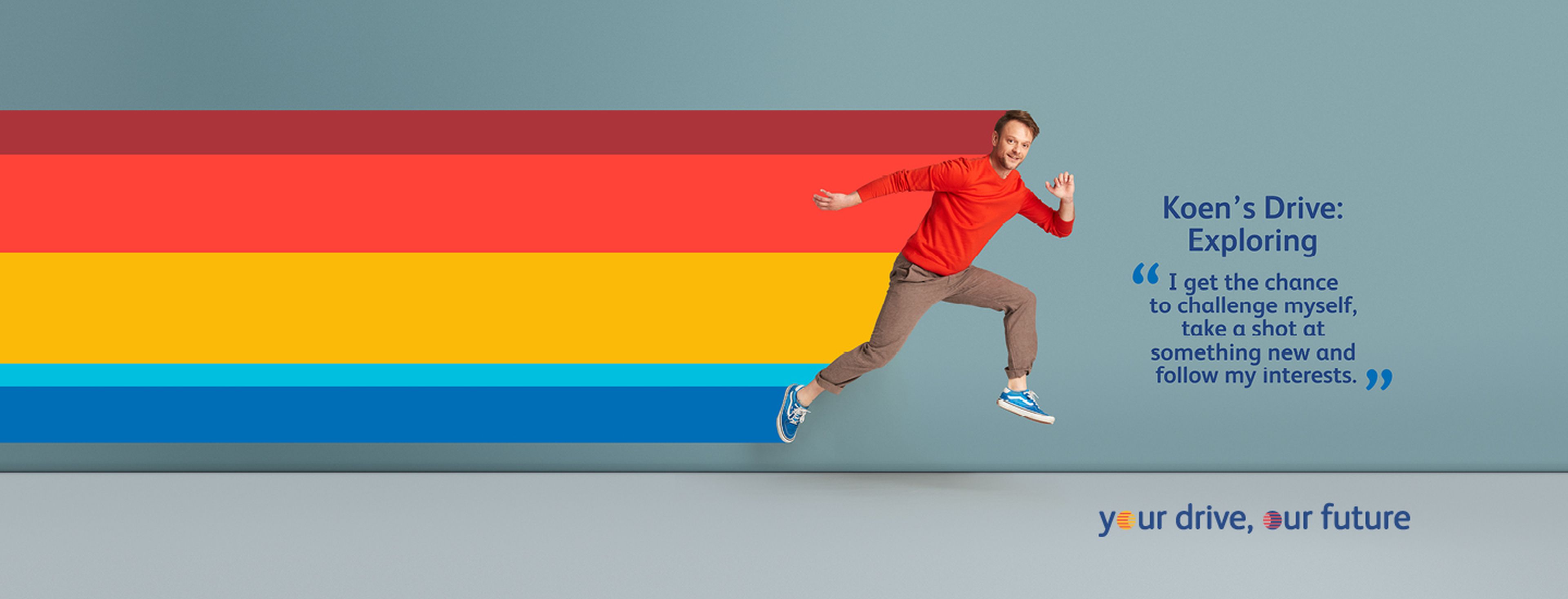 Een persoon springt energiek in de lucht tegen een effen blauwe achtergrond en draagt een felrode trui, opgerolde bruine broek en blauwe sneakers.