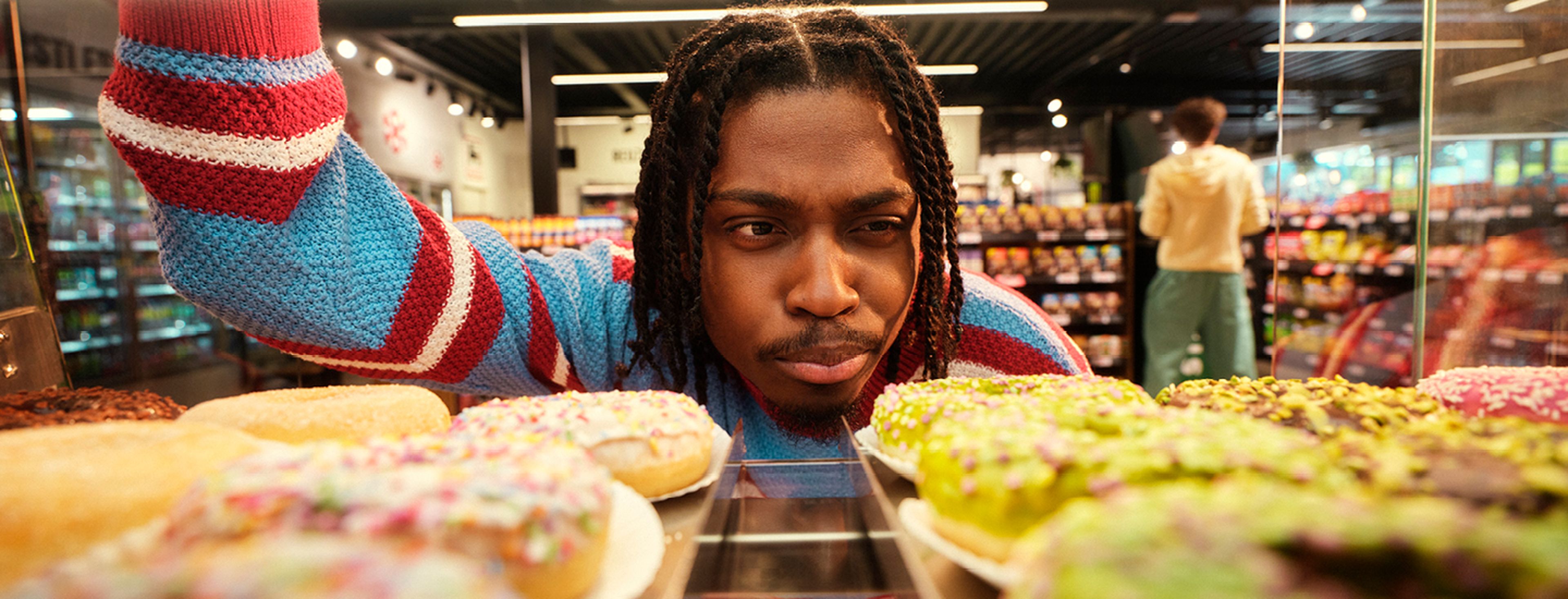 Personne avec des dreadlocks regarde des donuts en magasin ; autre personne à l’arrière-plan dans un rayon de snacks.
