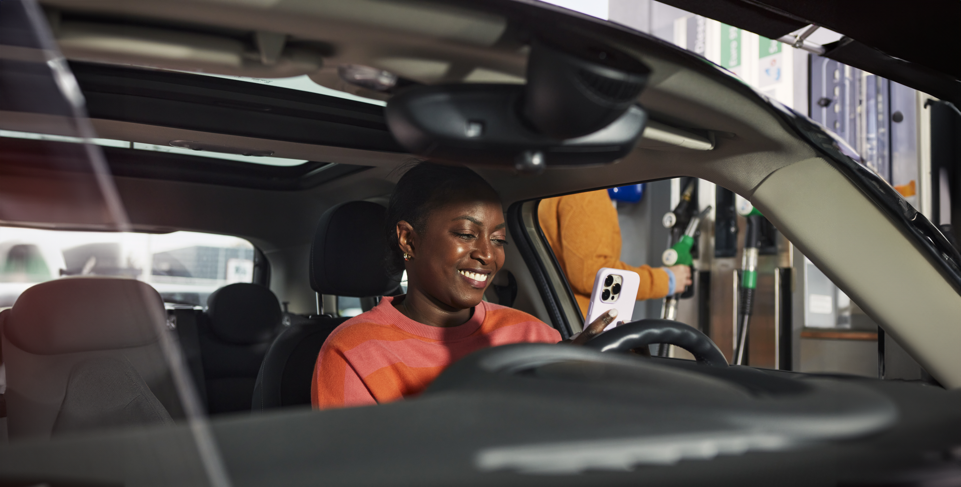 Woman in an orange sweater looks at her white smartphone while sitting in her car.