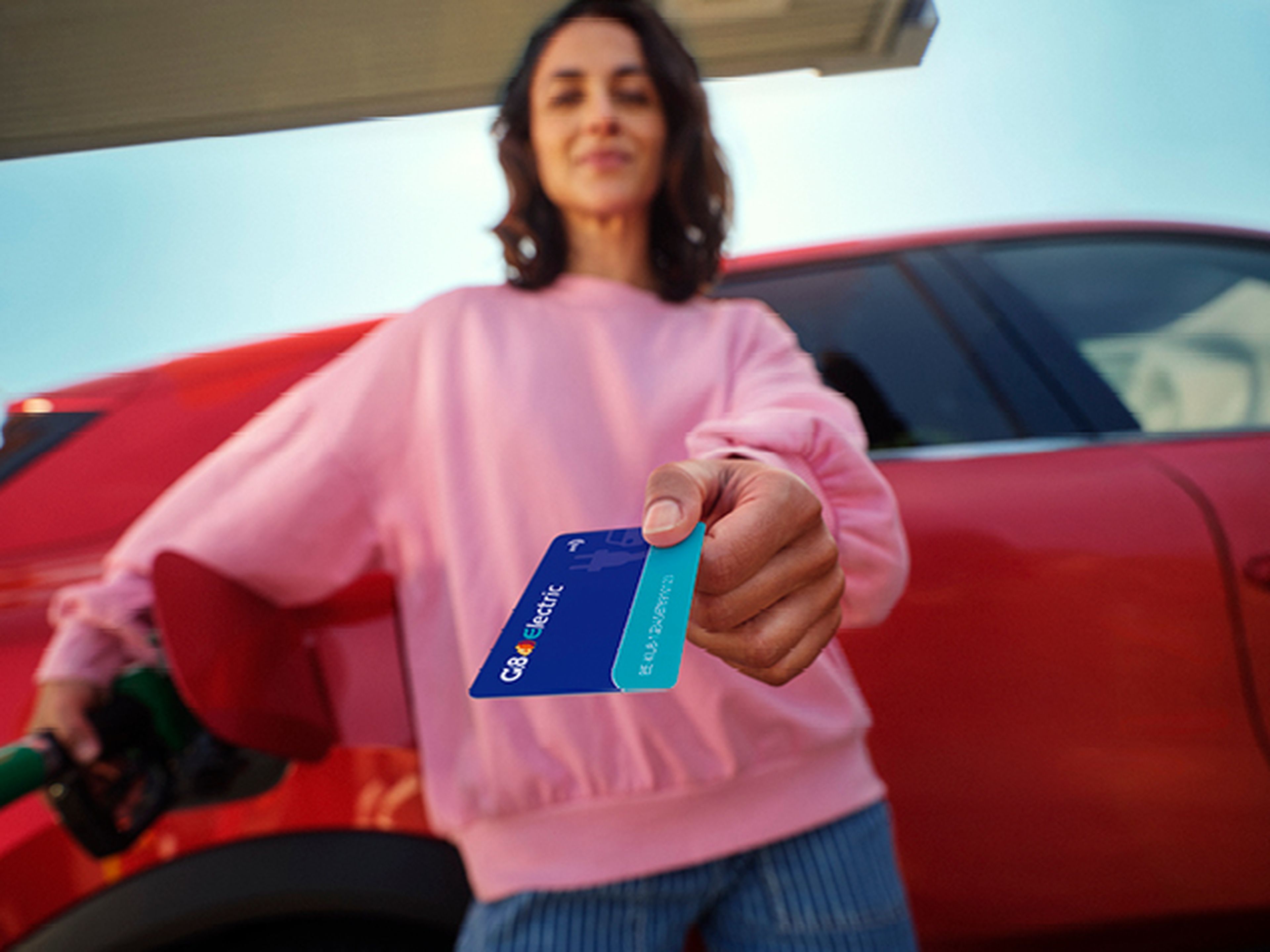 Een vrouw in een roze trui houdt een Q8 electric card naar de camera terwijl ze haar rode auto aan het laden is