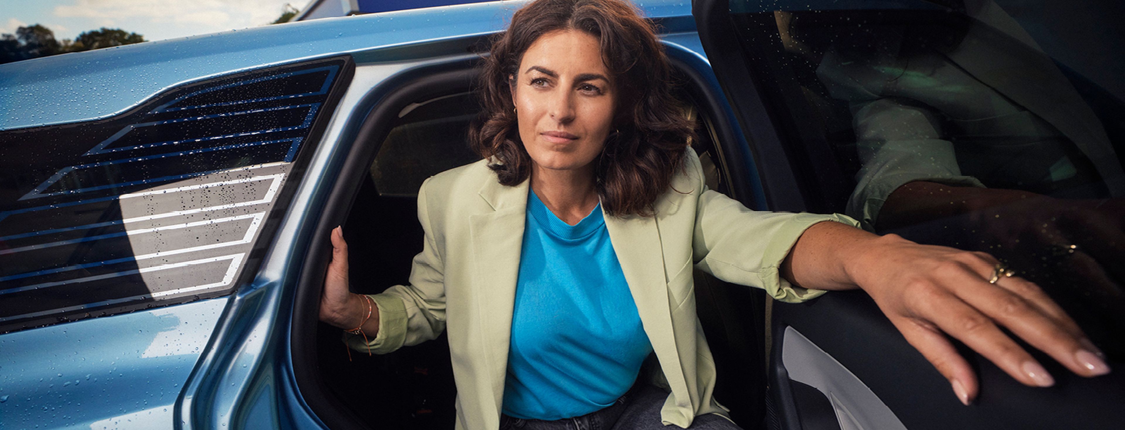 Een vrouw met donker krullend haar stapt uit een blauwe elektrische auto. Ze draagt een beige blazer, een felblauw T-shirt en een donkere jeans.