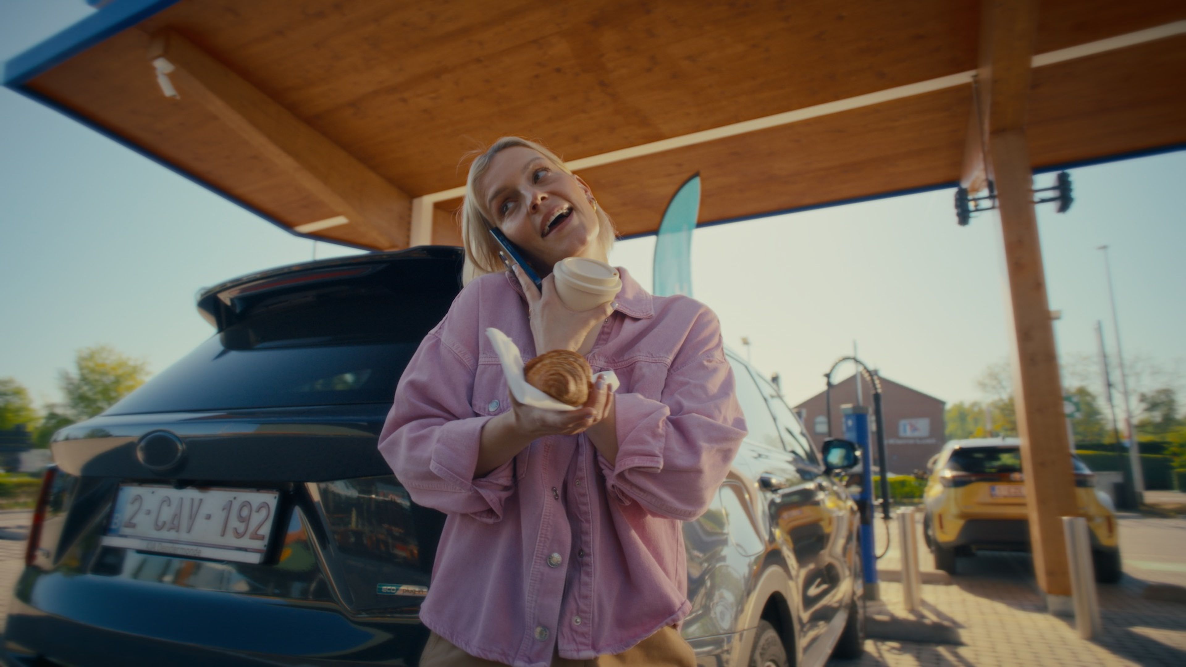 Femme en train de téléphoner, tenant un café et un croissant, pendant qu'elle recharge sa voiture.