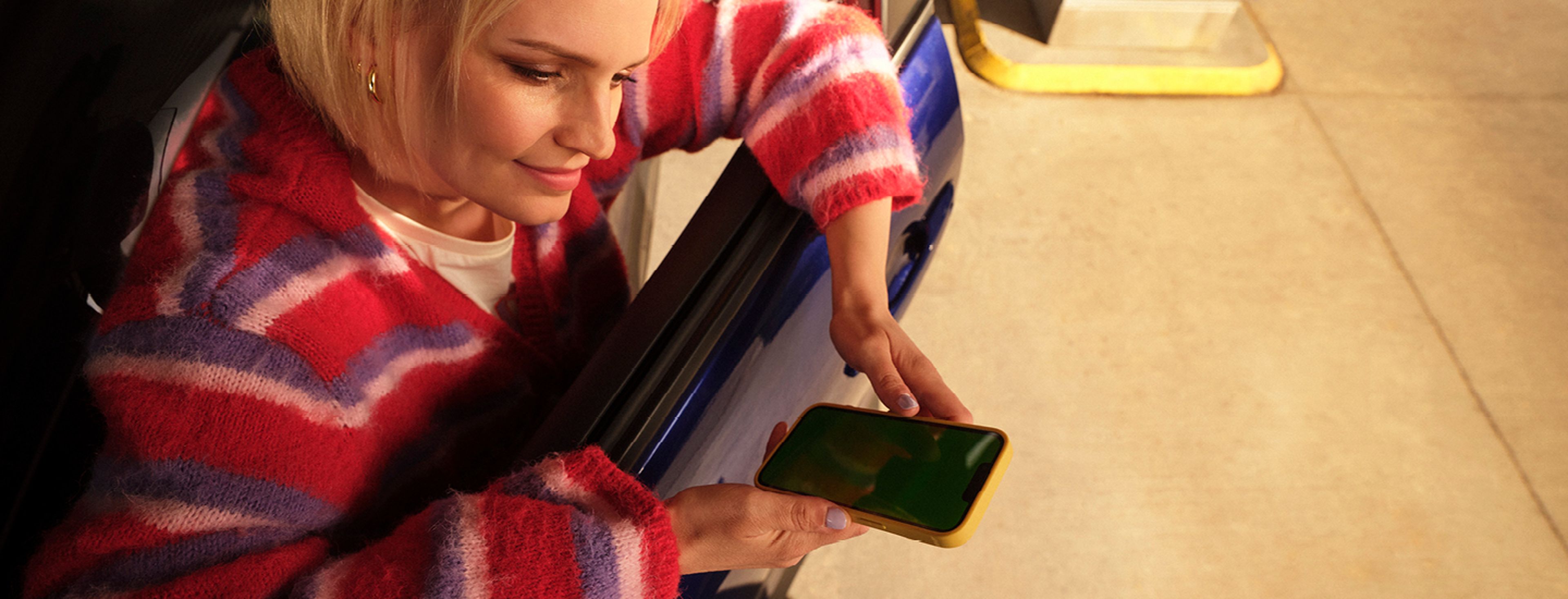 Een vrouw met blond haar en een kleurrijke gestreepte trui zit in haar auto bij een Q8 tankstation. Ze houdt haar smartphone uit het raam, klaar om iets te scannen of te betalen.
