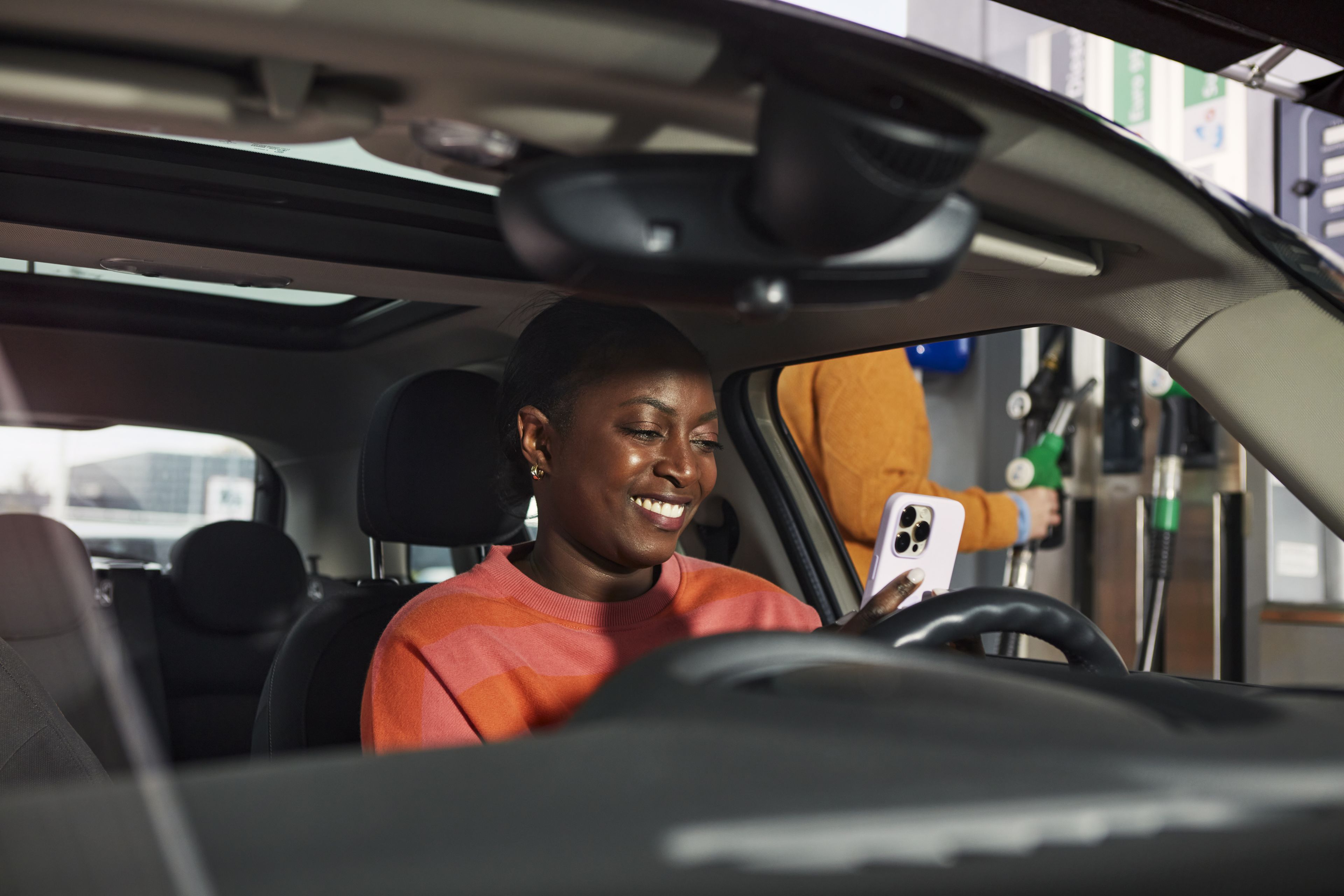 Vrouw met oranje trui kijkt op haar witte smartphone en zit in haar auto.