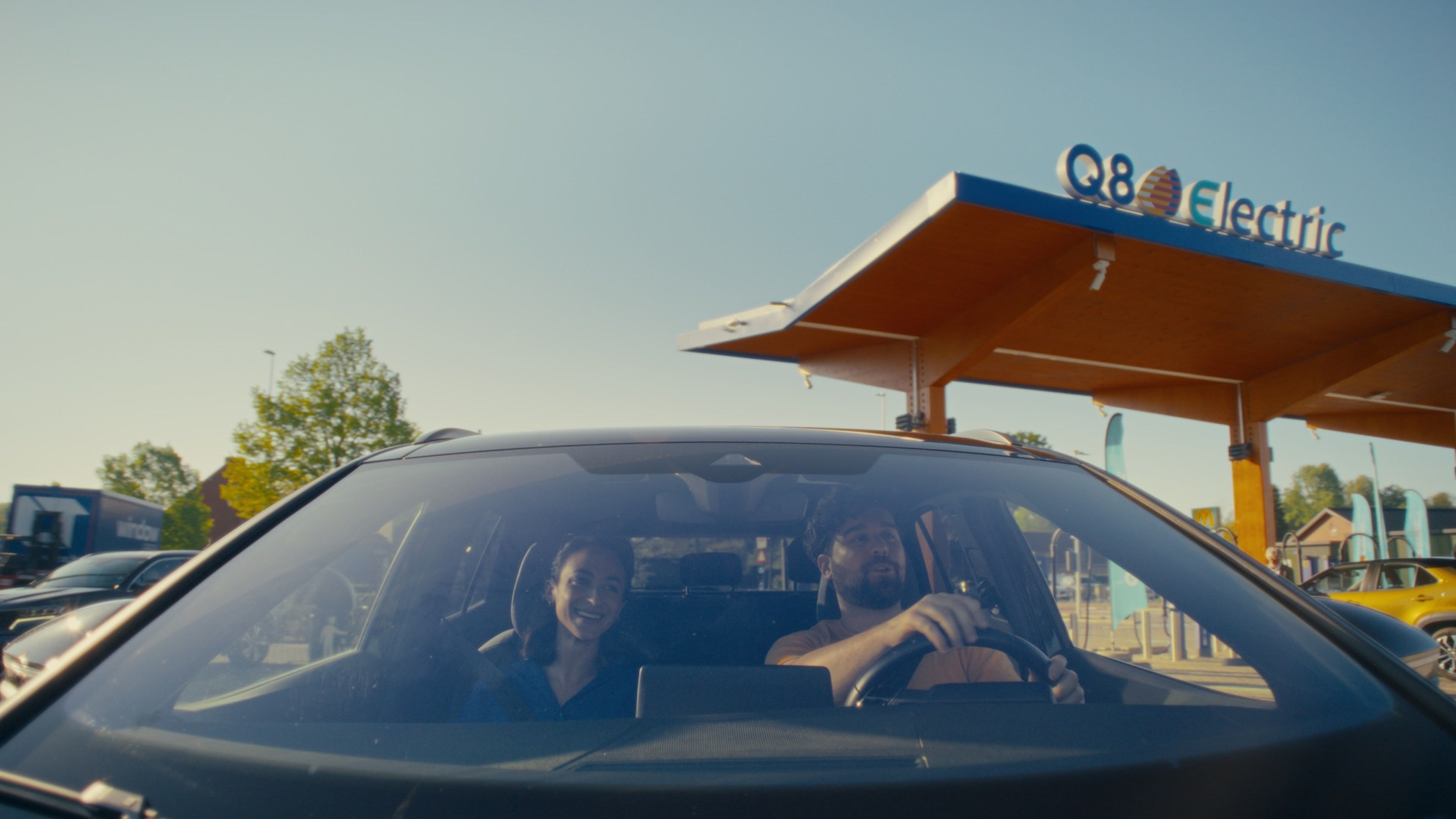Twee mensen in een elektrische wagen rijden weg van een Q8 electric-station, met het zonnige laadpunt en het herkenbare Q8 electric logo op de achtergrond.