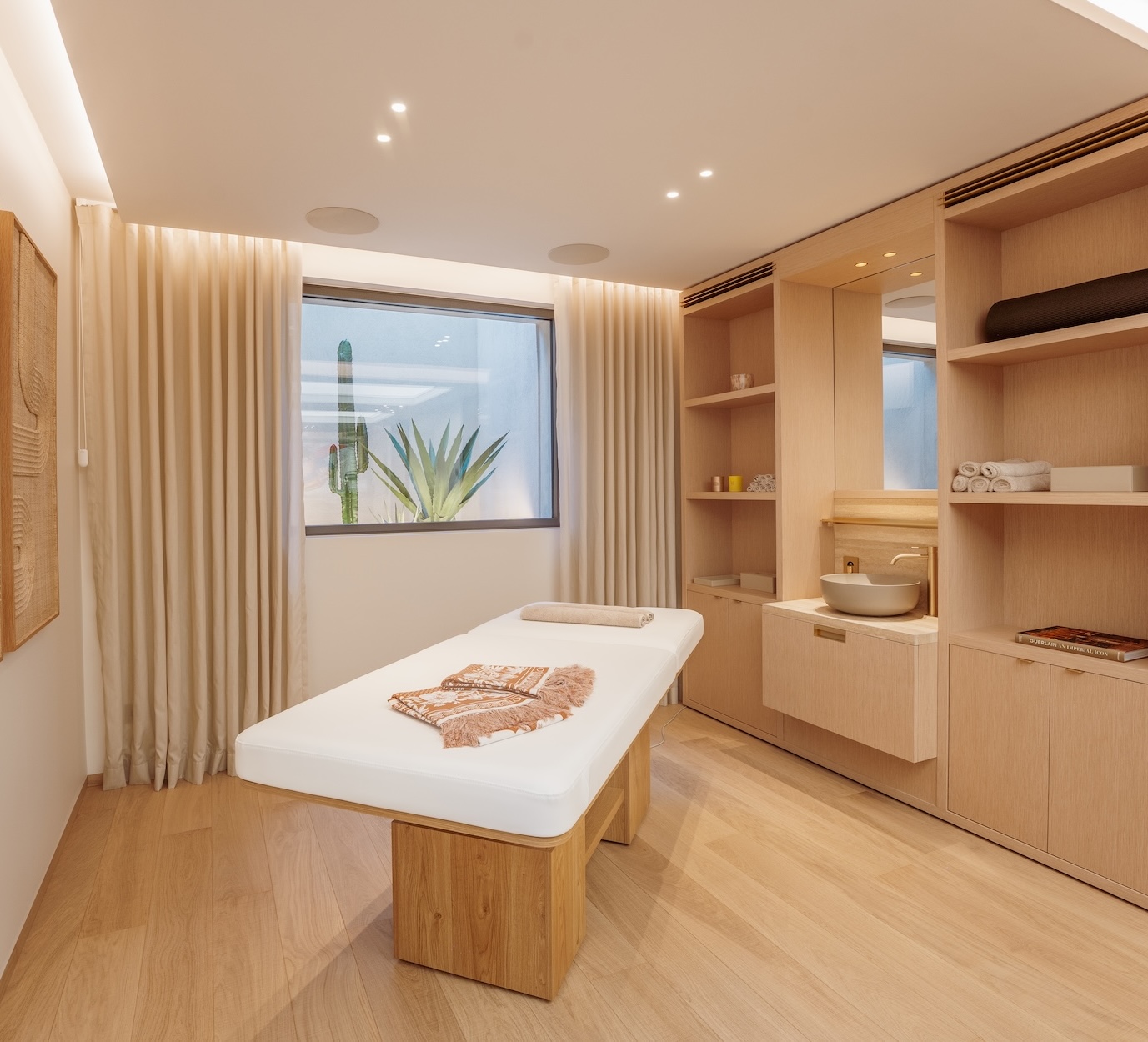 Modern massage room with a wooden treatment table, soft lighting, and a window view of plants. Shelves hold towels and spa essentials.