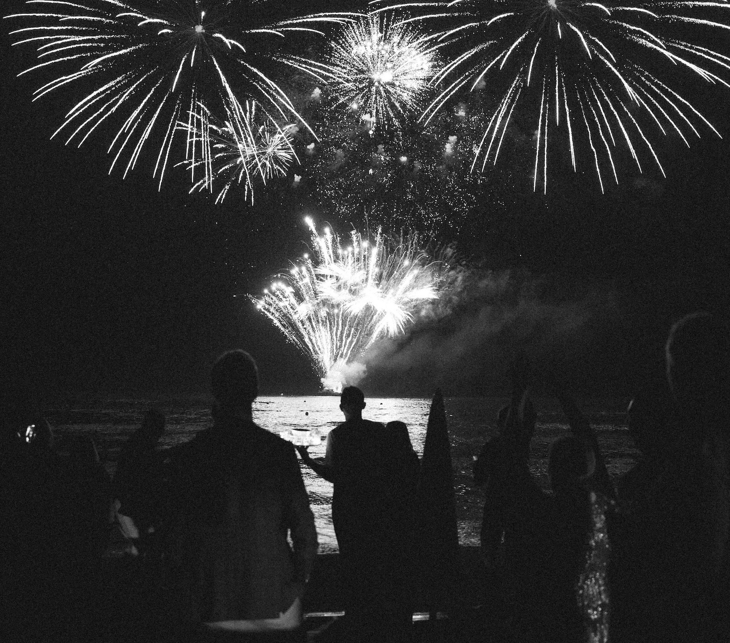 Silhouetted crowd watches fireworks display over water against night sky.