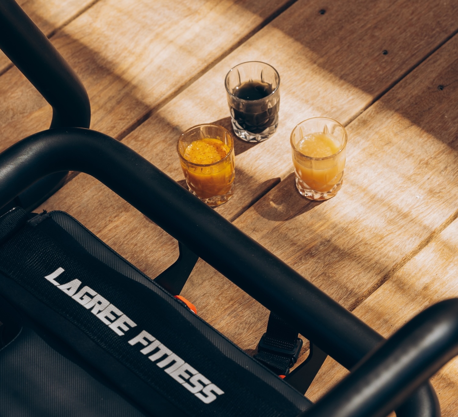 Three small glasses of dark and light drinks on a wooden surface next to a piece of Lagree Fitness equipment. Shadows cast across the scene.