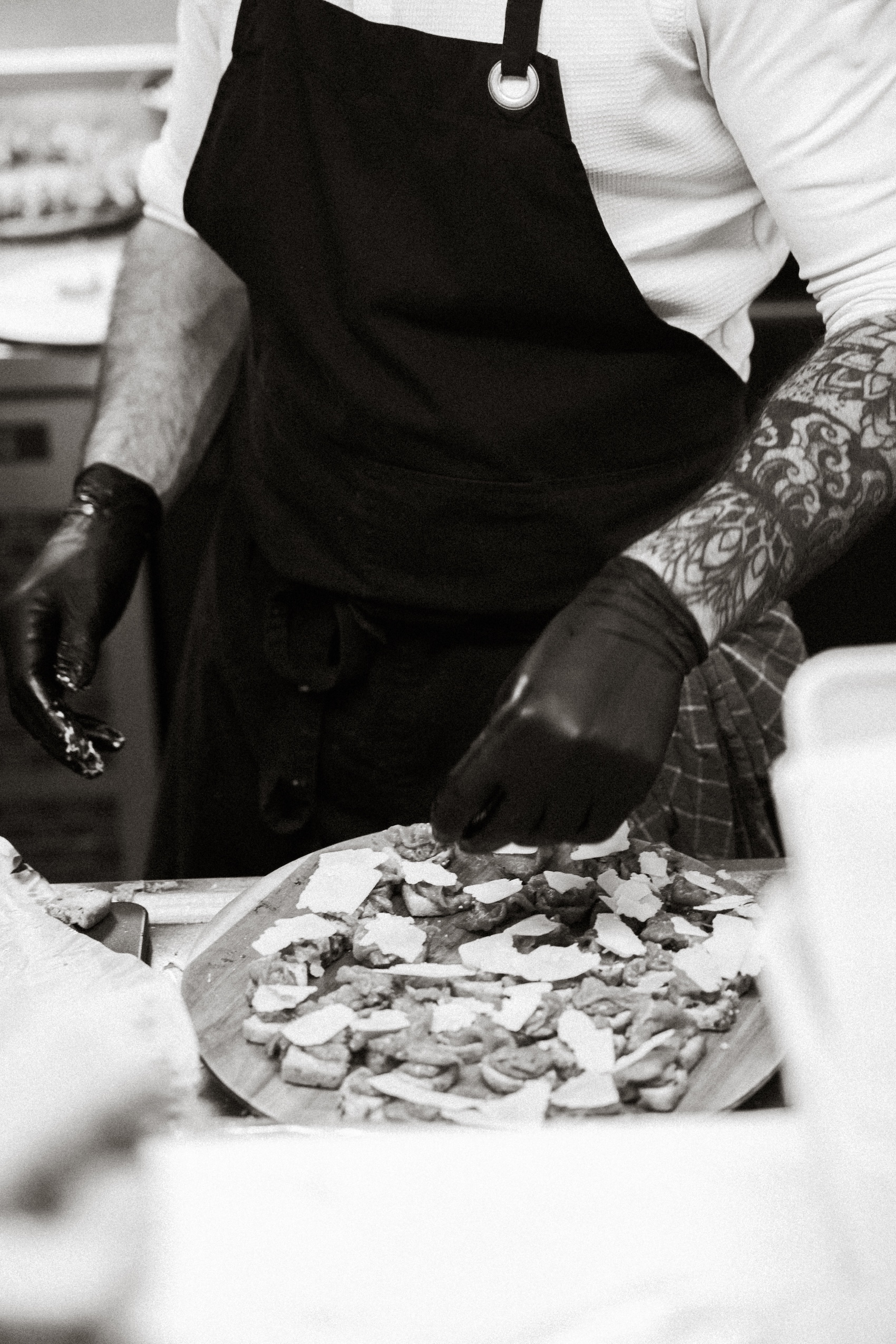 Chef with tattooed arms and gloves arranges toppings on a flatbread in a kitchen. Black and white image.
