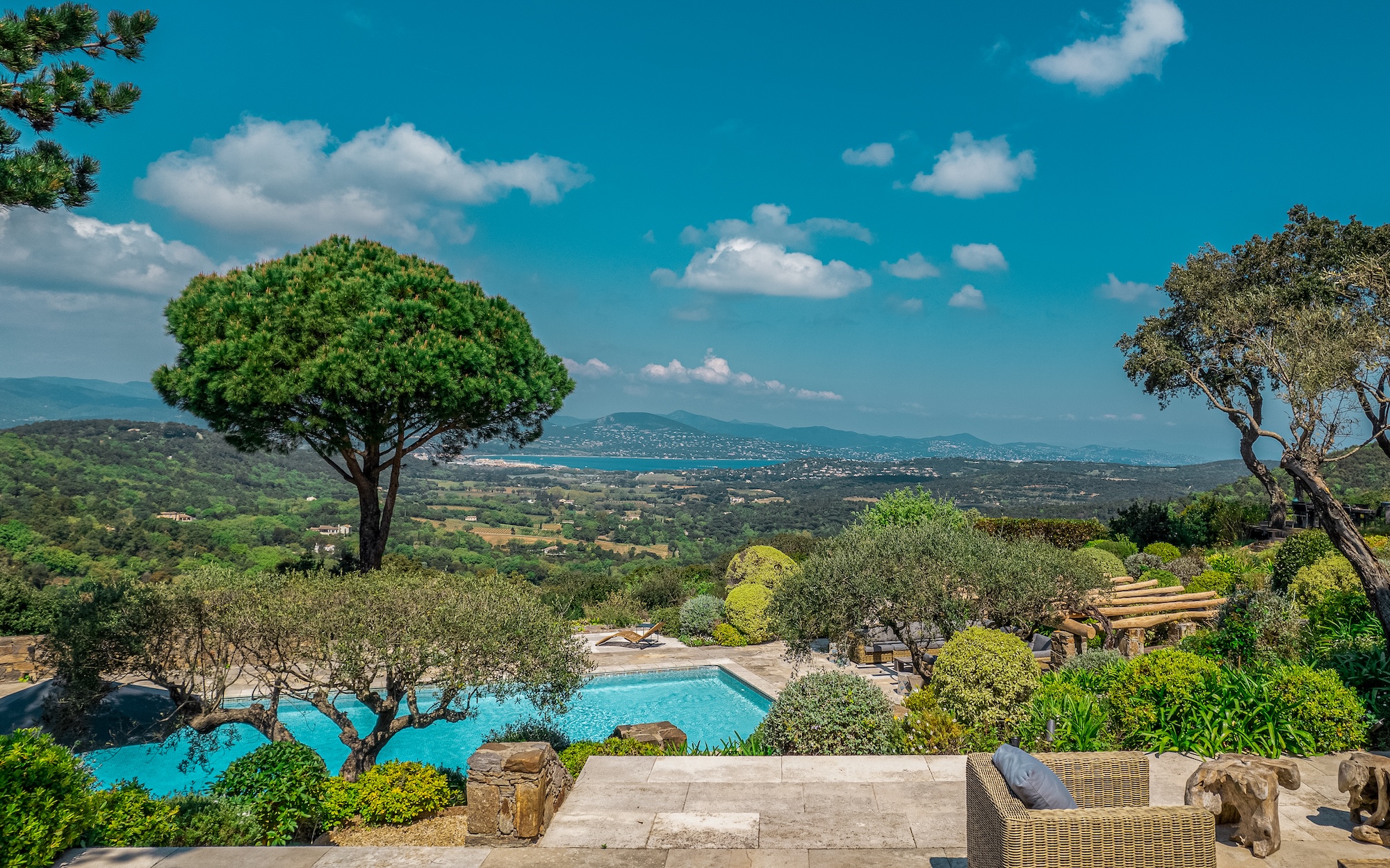 Scenic landscape with a pool, lush greenery, and a large tree, overlooking a valley and distant mountains under a blue sky with scattered clouds.