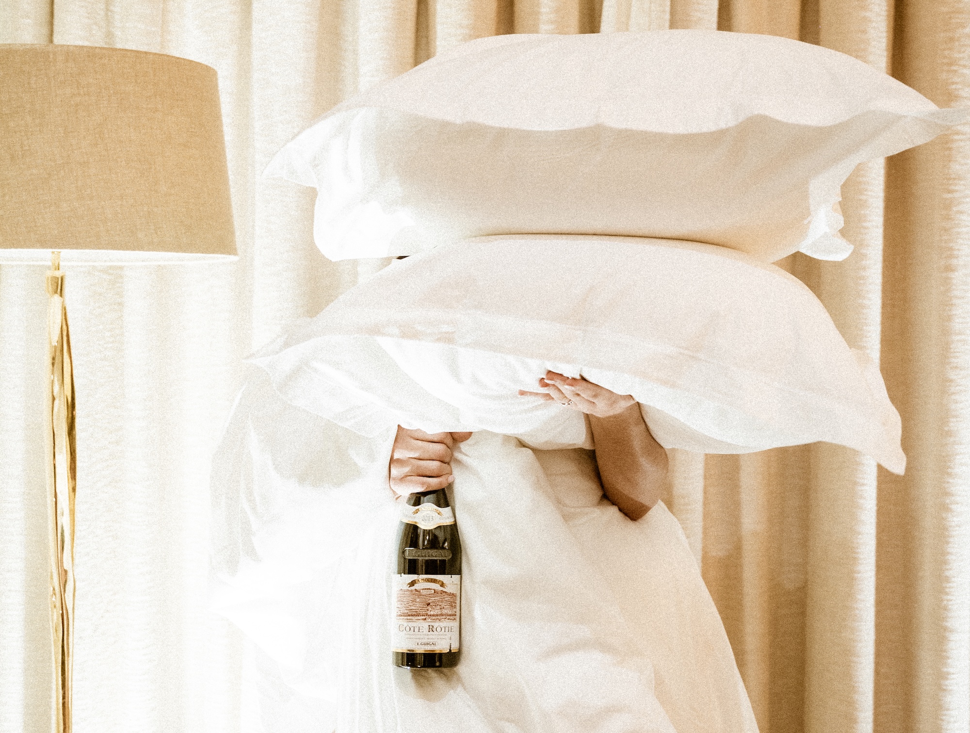 Person holding three pillows and a wine bottle, partially obscured, standing near a tall lamp with beige curtains in the background.