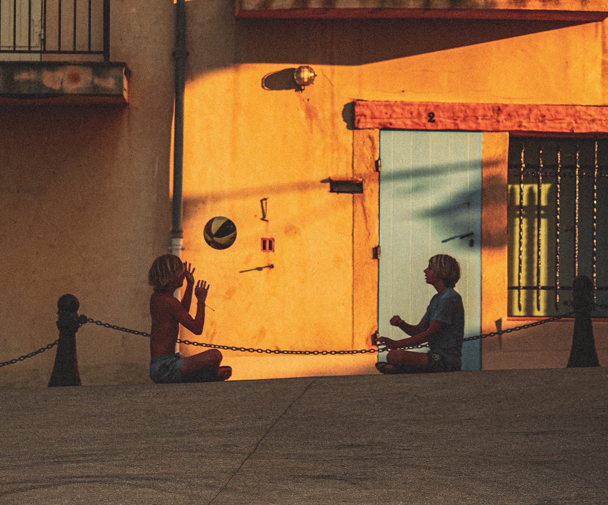 Two children sit on a sloped street playing with a ball. Warm sunlight casts long shadows on a wall with a blue door.