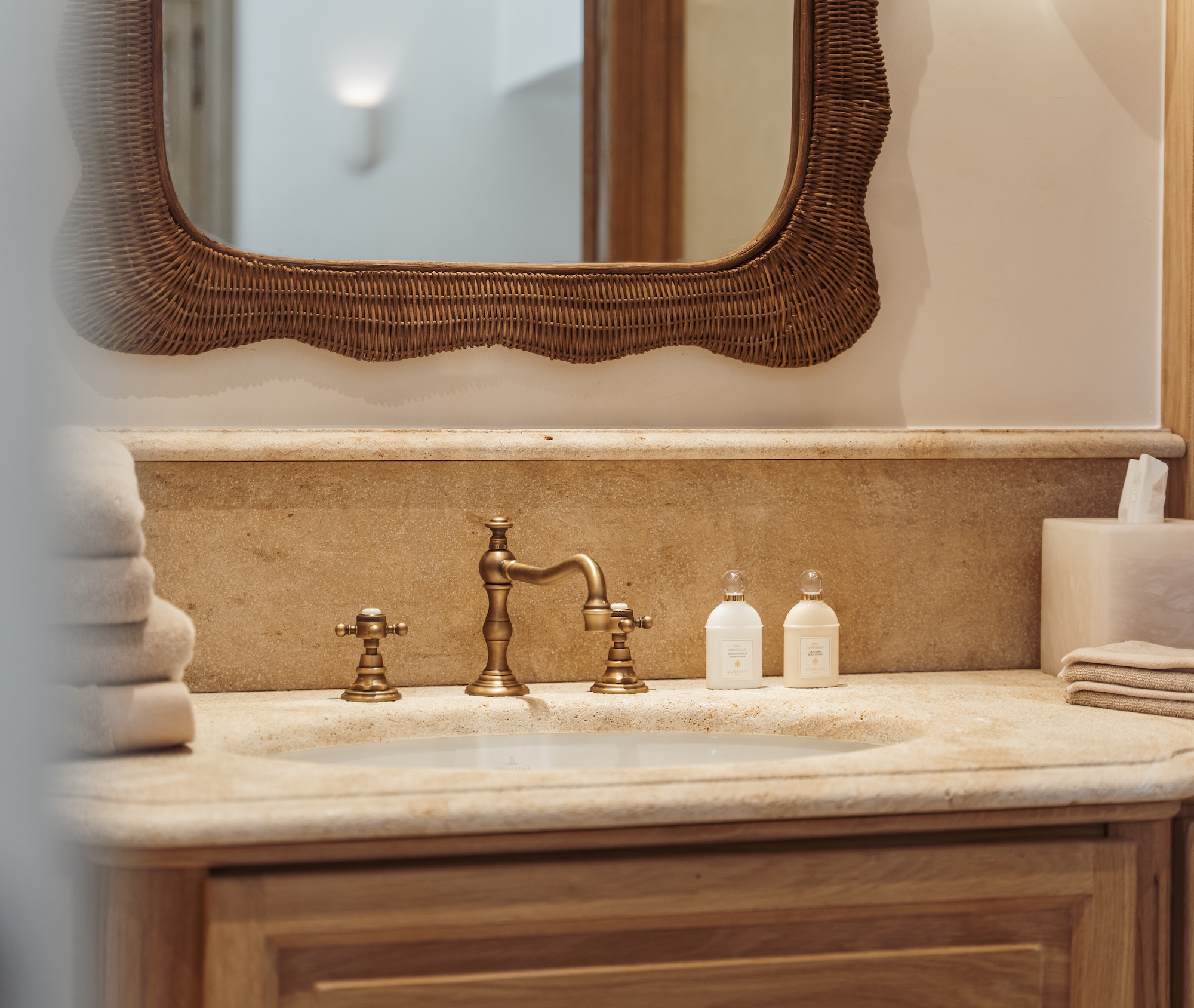Elegant bathroom sink with brass faucet, beige countertop, rectangular mirror, neatly folded towels, and small bottles of toiletries.