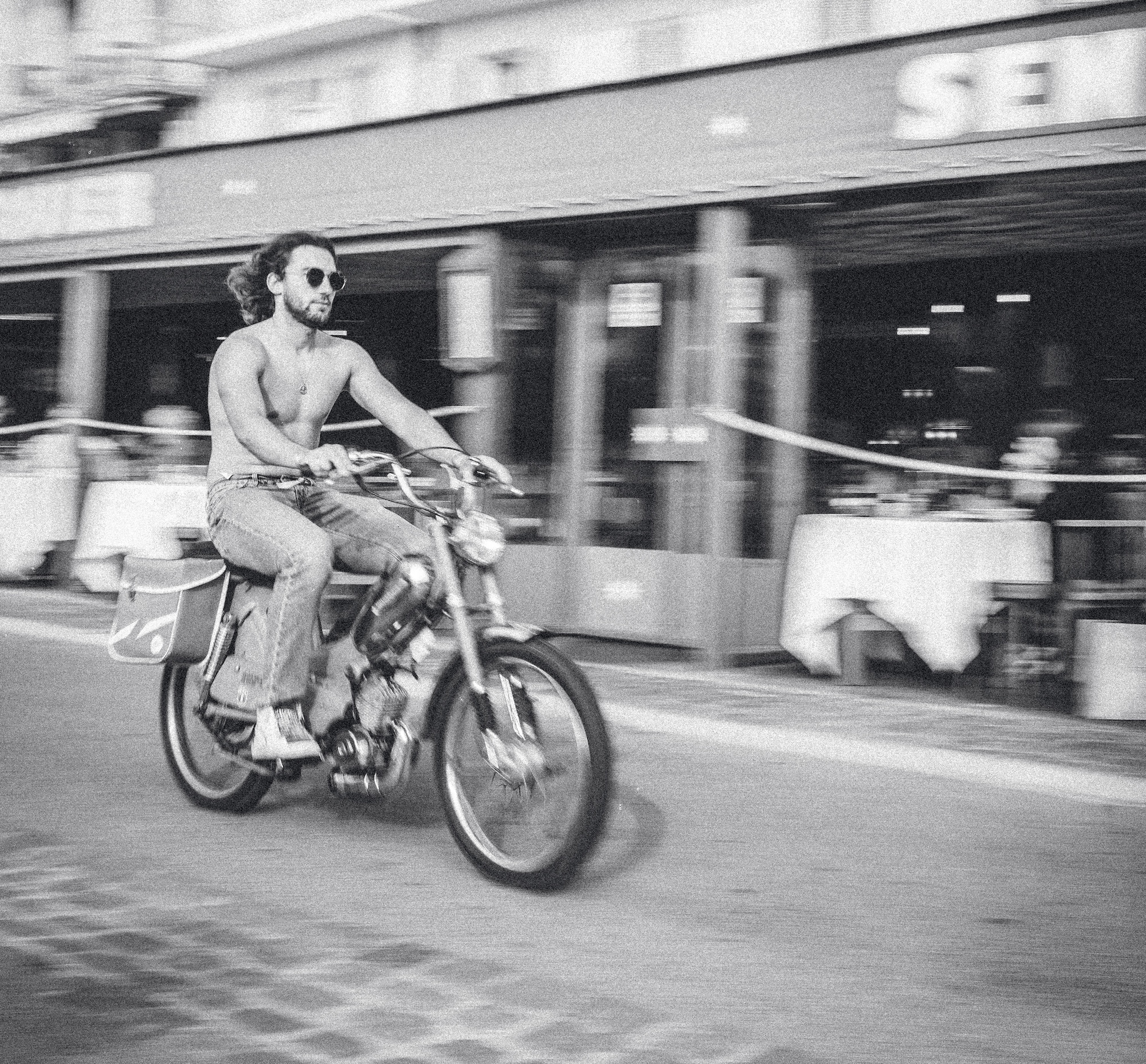 Shirtless man with sunglasses rides a vintage motorcycle quickly past a cafe, blurred background, black and white photo.