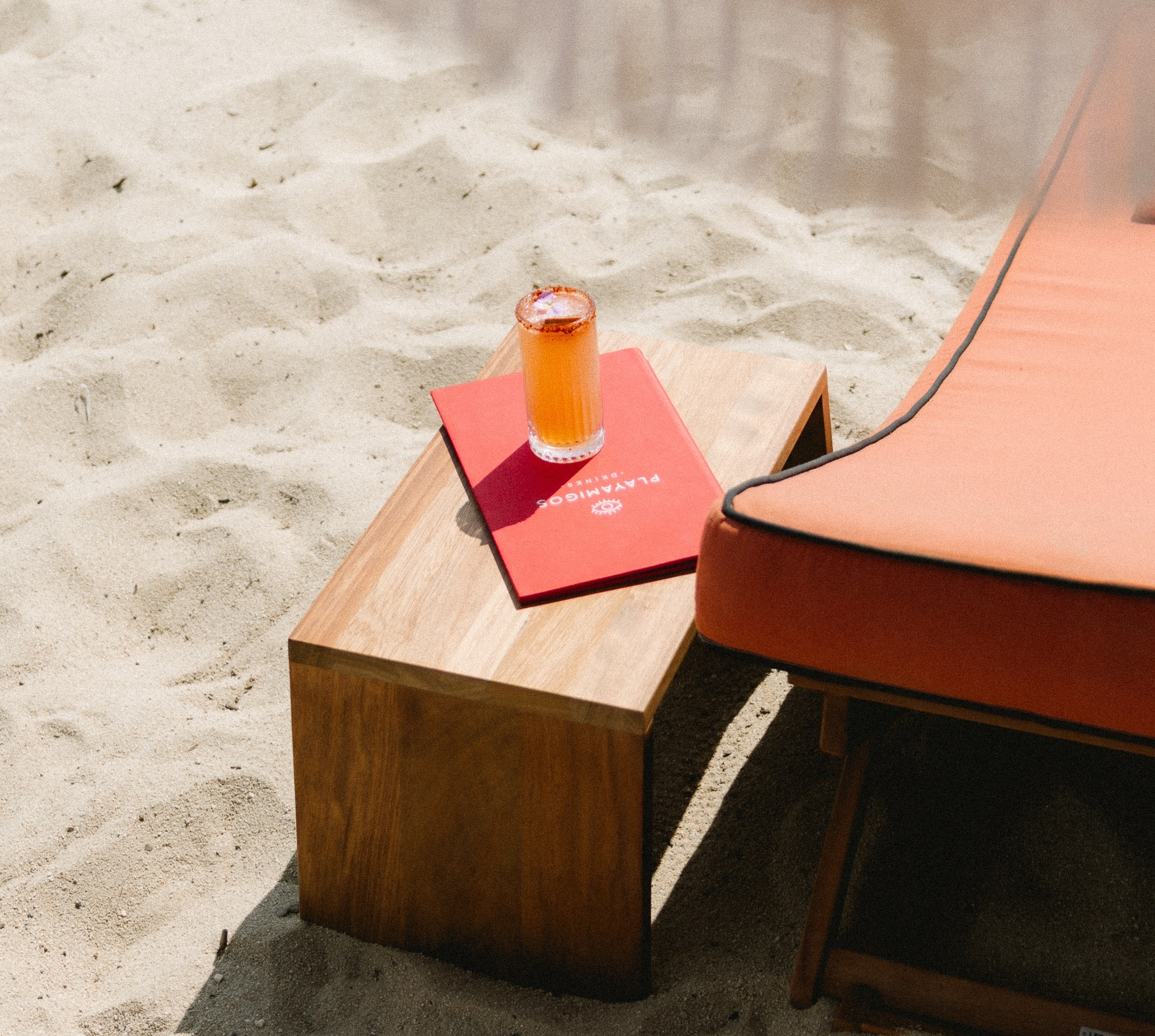 Beach scene with a wooden side table holding a red menu and a drink beside an orange cushioned lounge chair on the sand.