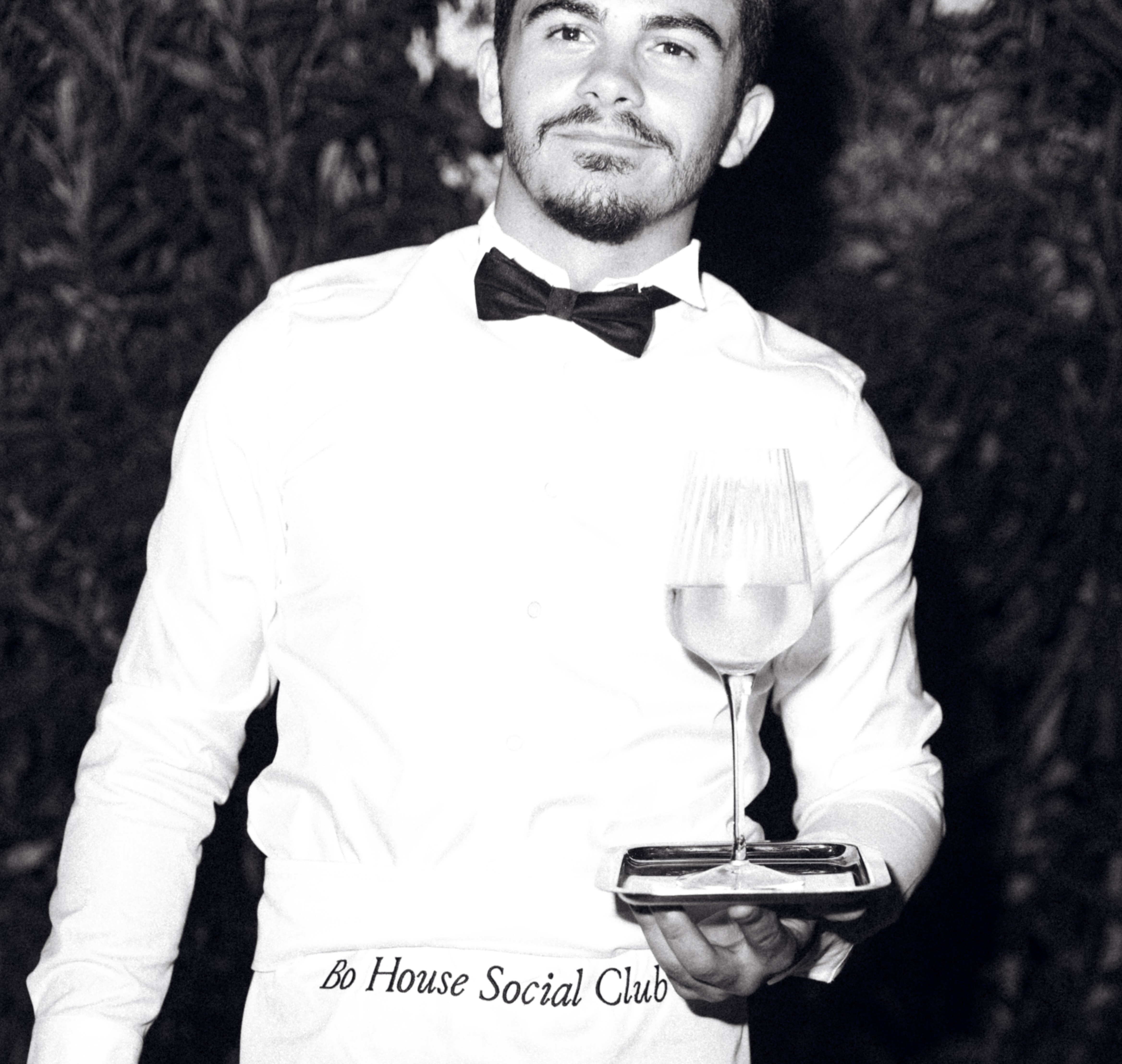 Waiter in a bow tie holding a tray with a glass of wine. His apron reads 