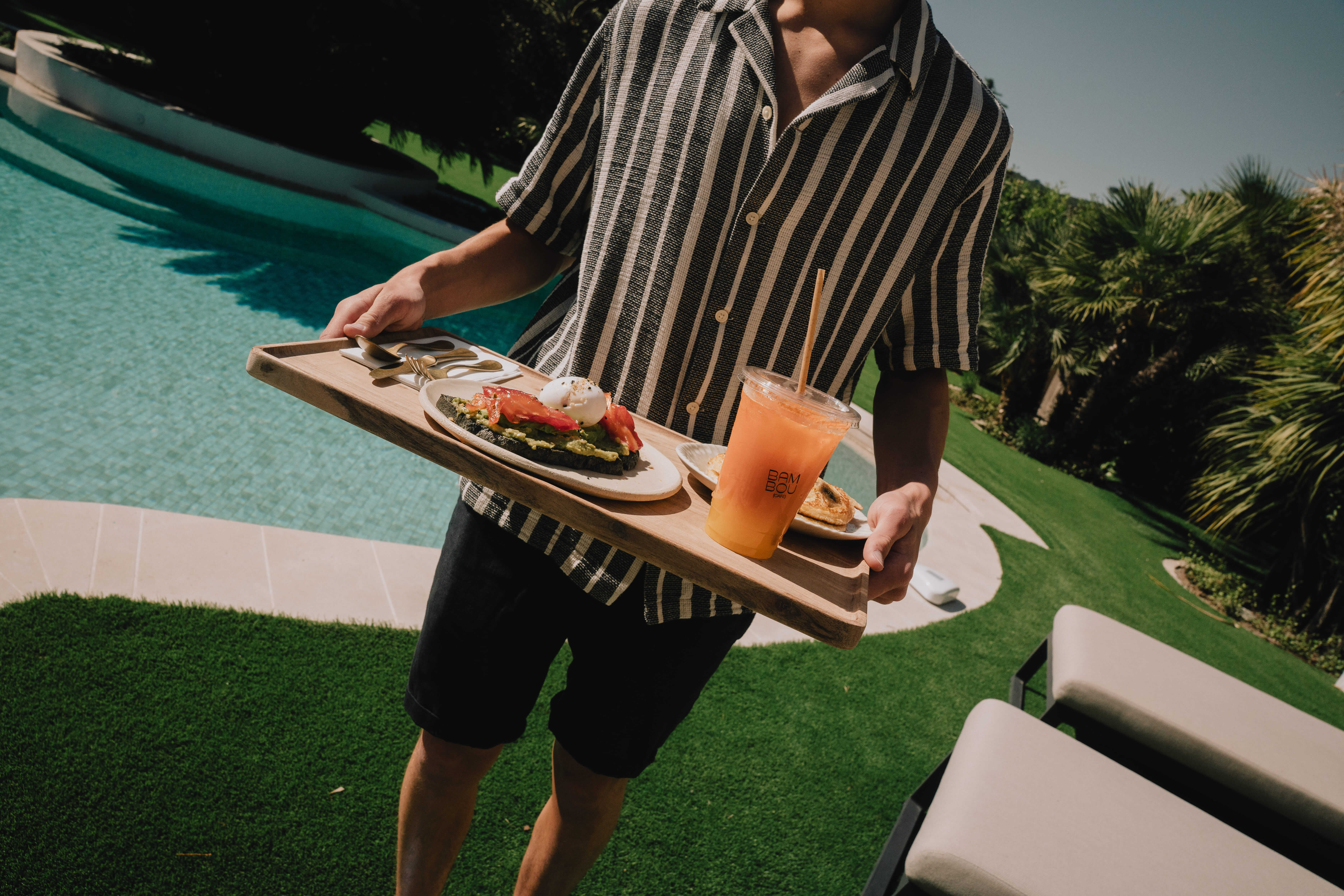 Person in striped shirt carrying a tray with avocado toast, poached eggs, and an orange drink by a pool, surrounded by greenery.