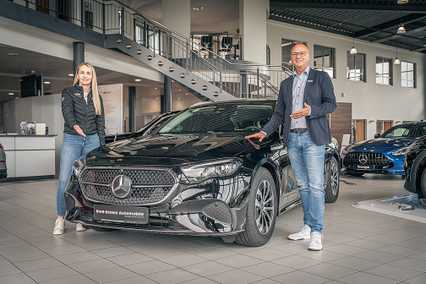 A man and woman stand beside a black Mercedes-Benz in a showroom, smiling and gesturing towards the car.
