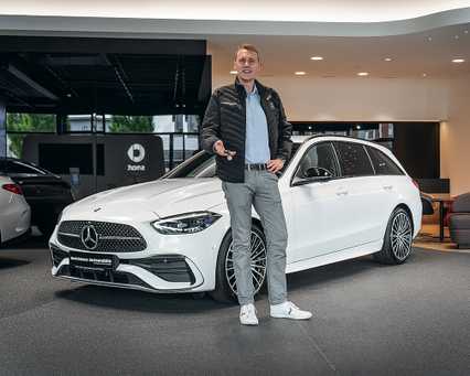A man stands beside a white Mercedes-Benz in a showroom, gesturing with his hand. He's wearing a black jacket, blue shirt, and gray pants.