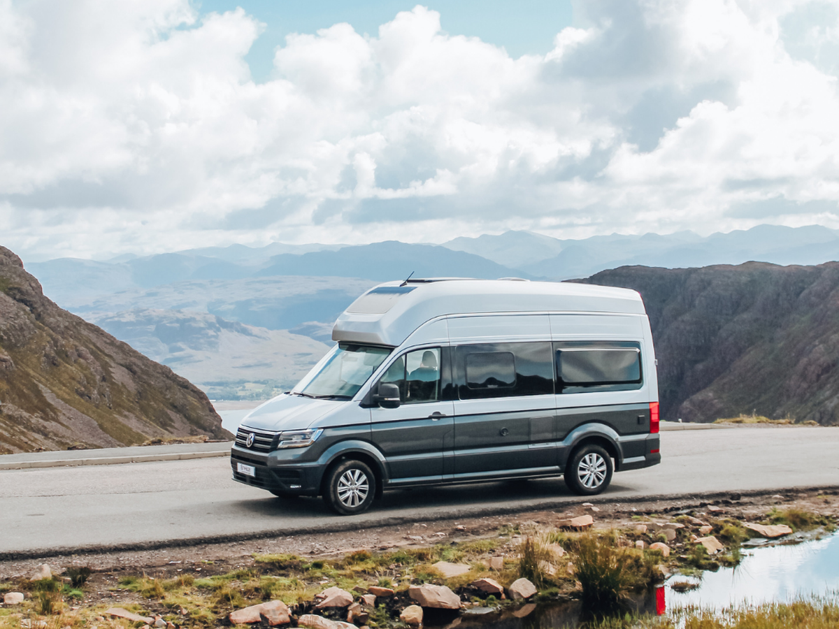 Camper van parked on a scenic mountain road, surrounded by rugged terrain and a cloudy sky reflected in a nearby puddle.