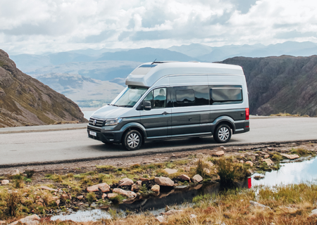 Camper van parked on a scenic mountain road, surrounded by rugged terrain and a cloudy sky reflected in a nearby puddle.