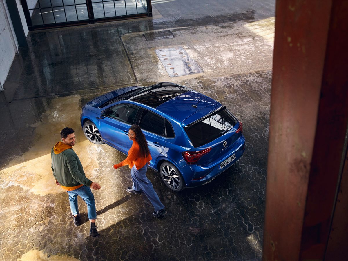 A man and woman walking near a parked blue car inside an industrial-style building with wet, textured flooring.