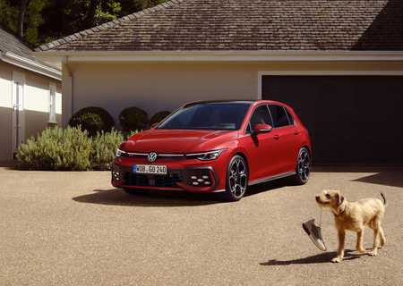 A red Volkswagen Golf 8 GTI hatchback parked in front of a garage, with a dog holding a shoe in its mouth walking nearby.