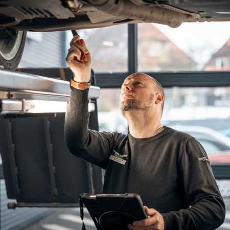 Mechanic inspecting a car's underside with a flashlight, holding a diagnostic tablet. He's wearing a dark long-sleeve shirt in a garage.