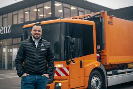 Man in a black jacket stands smiling in front of an orange garbage truck near a building with large windows.