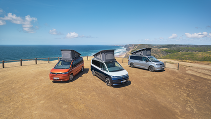 Three Volkswagen camper vans with popped roofs parked on a coastal cliff, overlooking the ocean and distant cliffs under a clear blue sky.