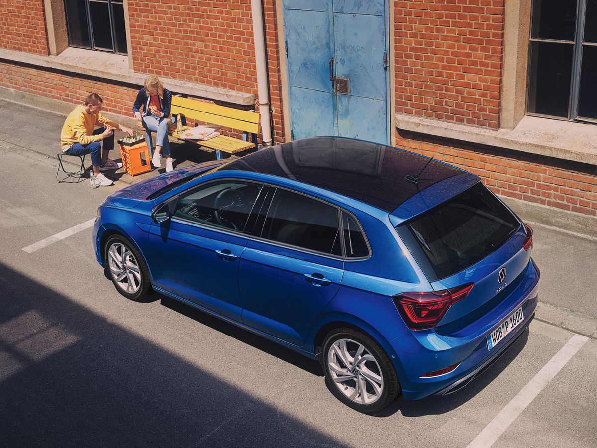 A blue polo hatchback car parked beside a brick building, with two people sitting on a yellow bench near a blue door, eating and chatting.