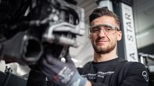 Mechanic wearing safety glasses and gloves, smiling while working on a car part in a garage setting.