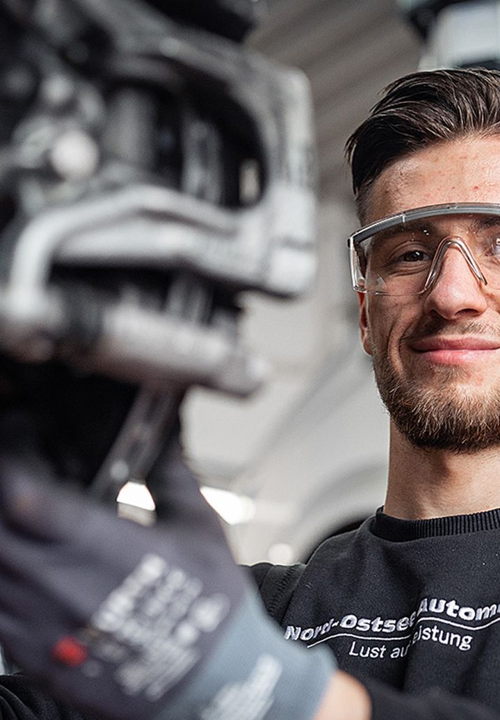 Mechanic wearing safety glasses and gloves, smiling while working on a car part in a garage setting.
