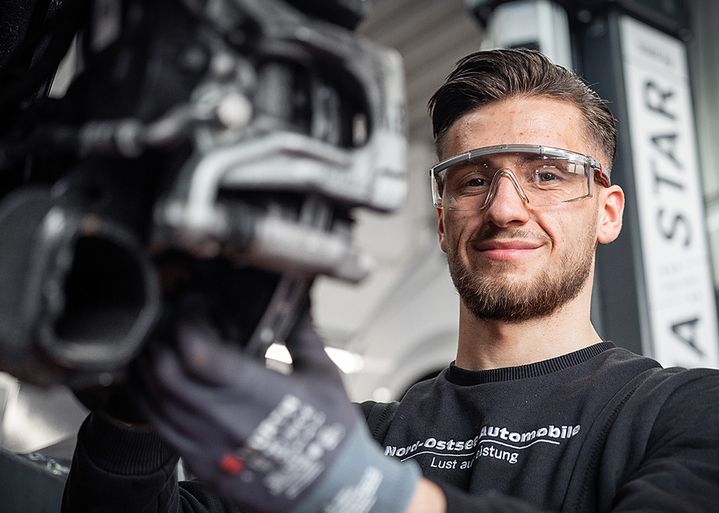 Mechanic wearing safety glasses and gloves, smiling while working on a car part in a garage setting.