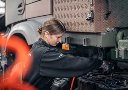 A person in a black jacket works on the engine of a large vehicle, focusing intently.